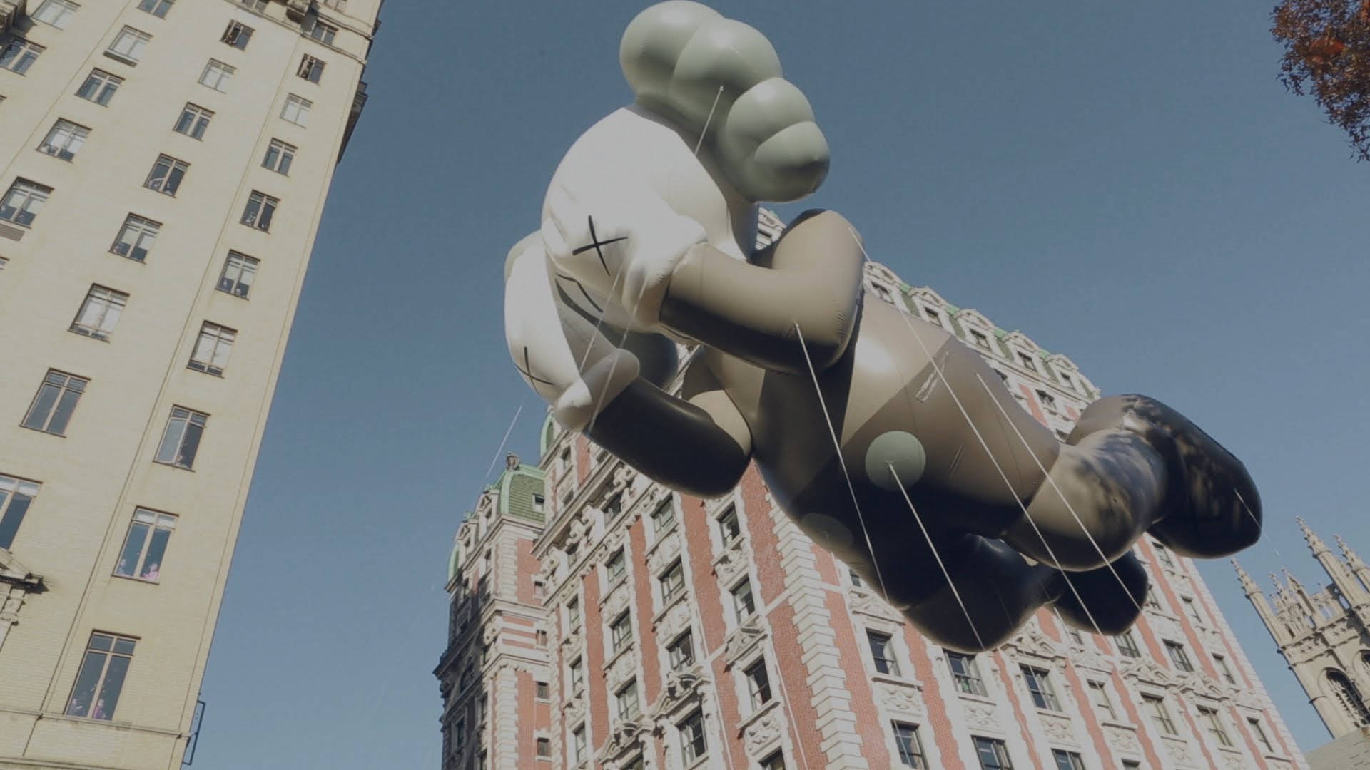 KAWS 桌布 壁紙 — 一個巨大的充氣KAWS熊在晴朗的藍天下漂浮在華麗的城市建築上方。