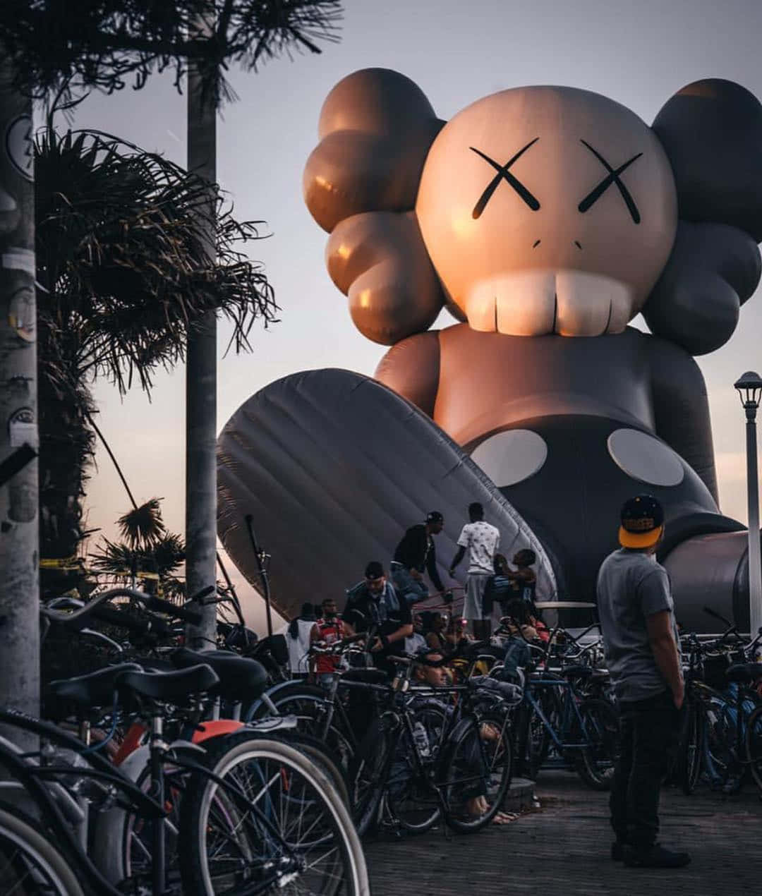 Kaws Holiday 桌布 壁紙 — 一個巨大的充氣KAWS角色在黃昏時分聳立於人群和腳踏車之上，背景有棕櫚樹。