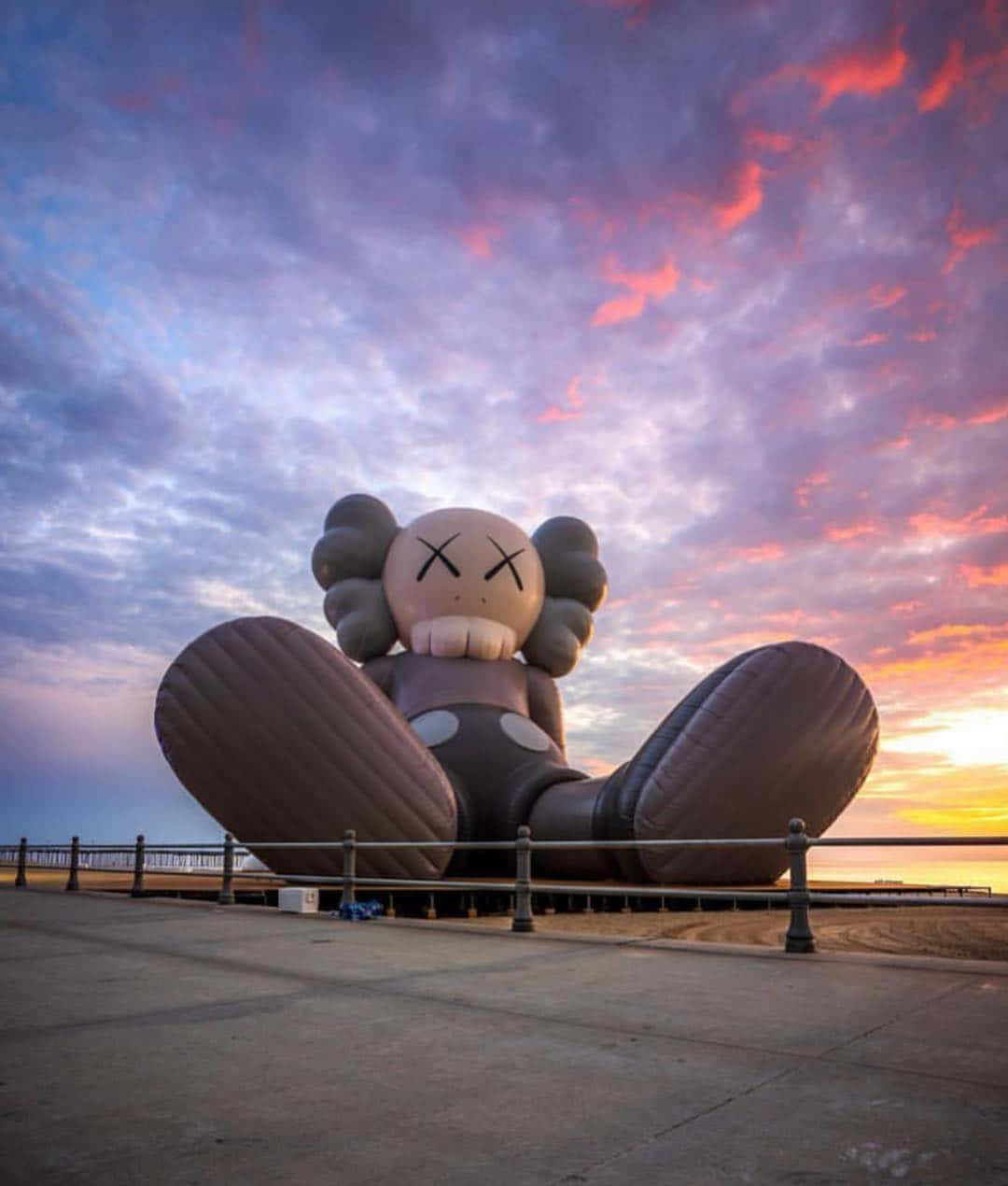 Kaws Holiday 桌布 壁紙 — 一個巨大的充氣KAWS雕塑盤腿坐在海邊長廊上，背景是戲劇性的日落天空。