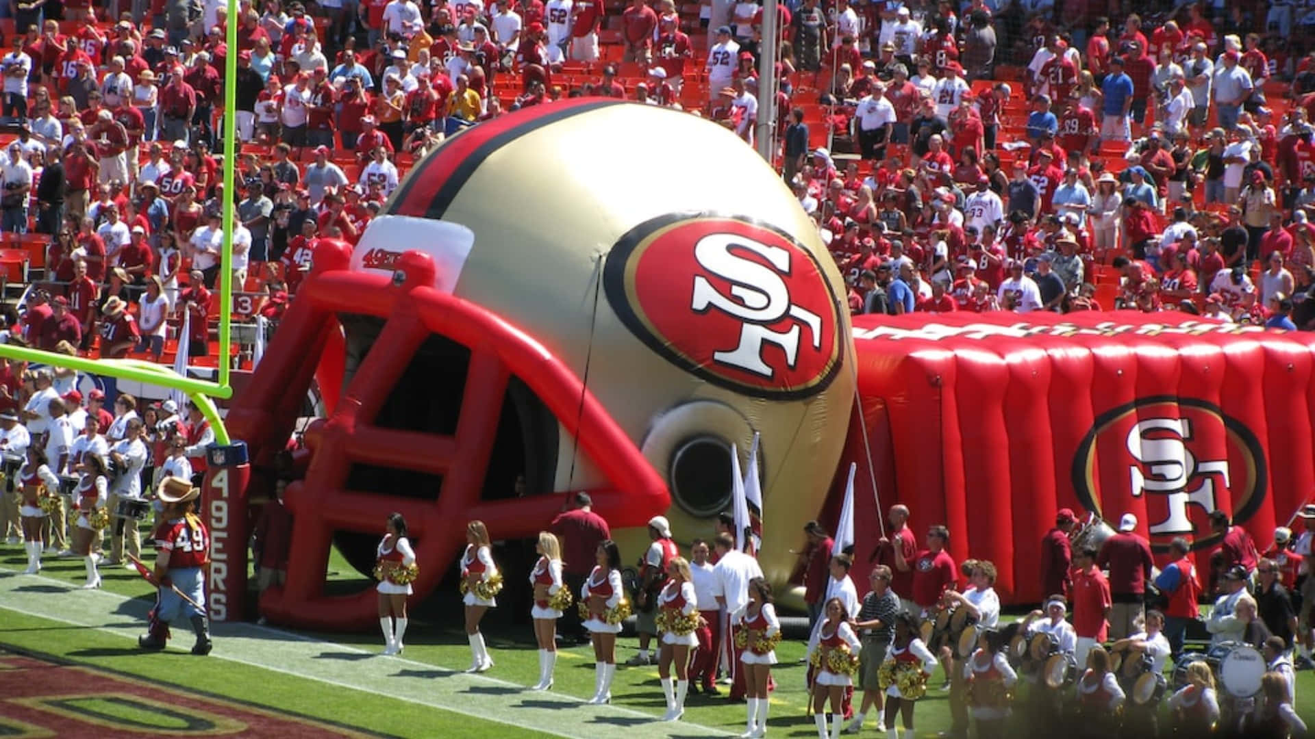 49ers 桌布 壁紙 — 一個巨大的充氣舊金山49人美式足球頭盔在陽光明媚的球場上佔據主導地位，啦啦隊在擠滿觀眾面前表演。