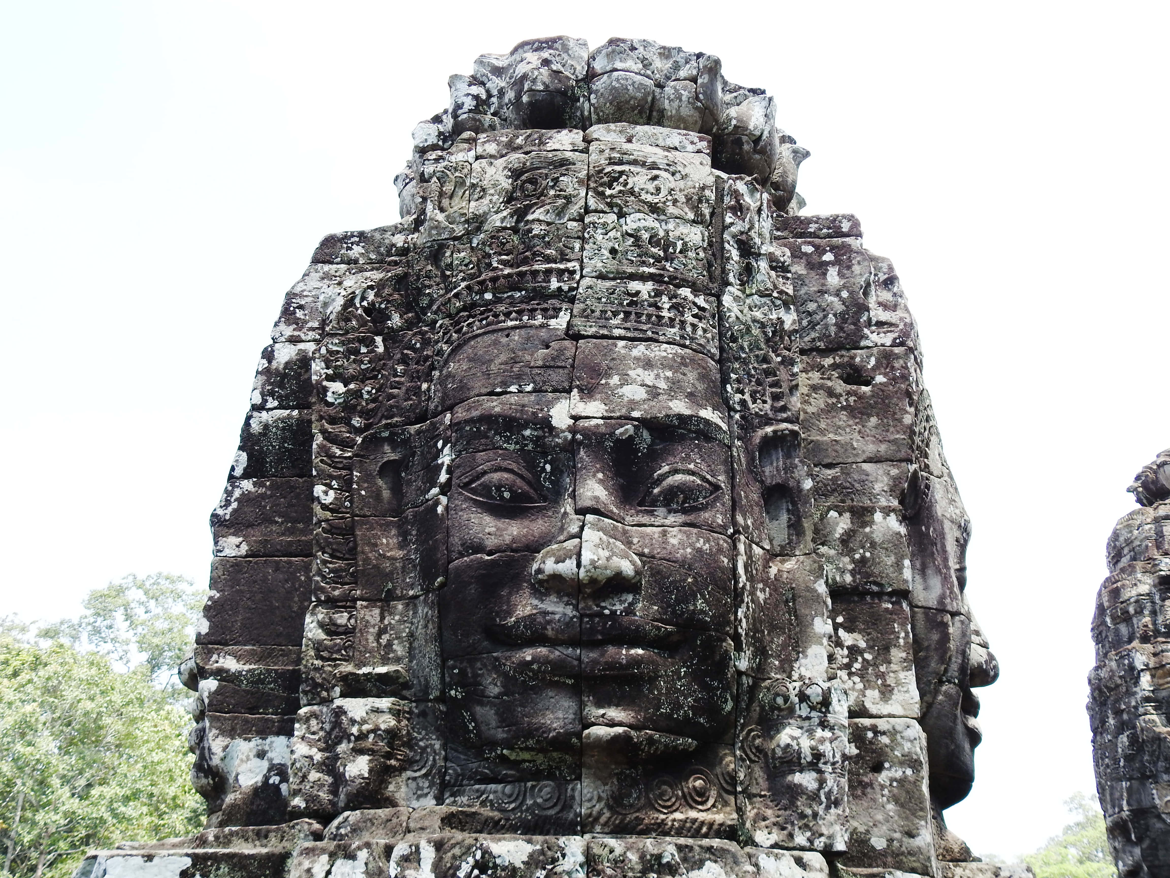 Cambodia 桌布 壁紙 — 一個巨大的古老石臉雕刻在寺廟塔樓上，帶有苔蘚質地和寧靜的表情，在陰天之下。