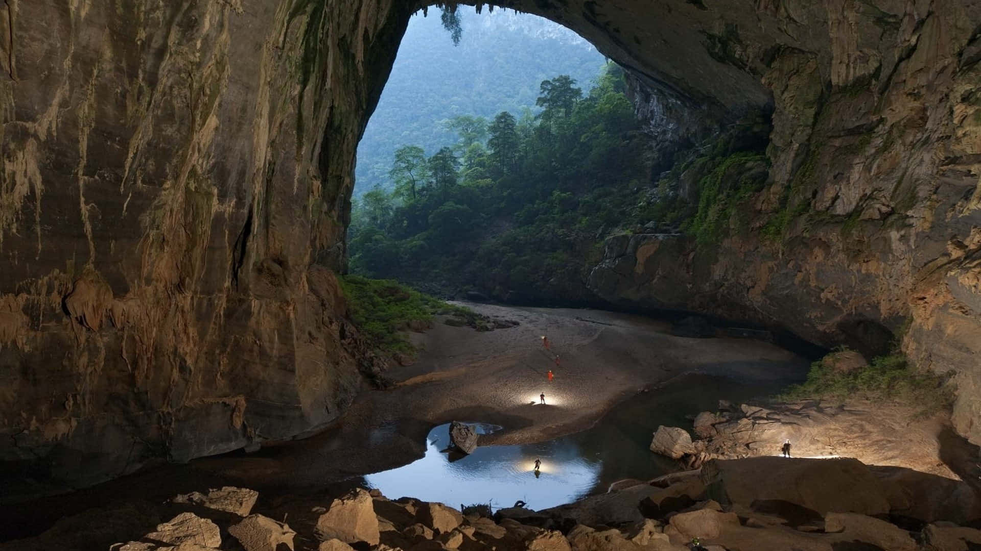 Cave 桌布 壁紙 — 一個巨大的天然洞穴通向遠處鬱鬱蔥蔥的森林，探險者們的頭燈照亮了岩石河床。