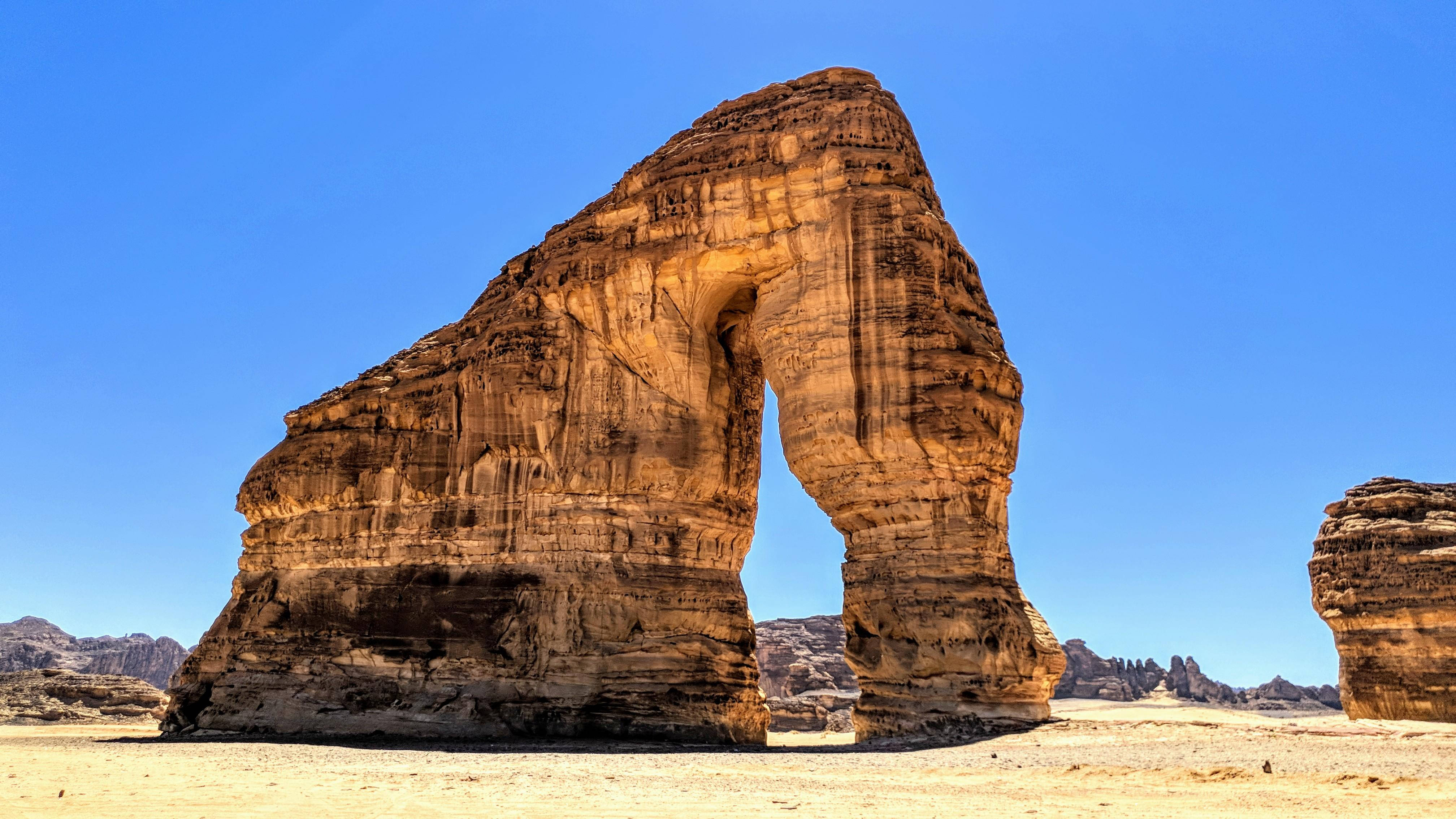 Saudi 桌布 壁紙 — 一個巨大的天然砂岩拱門在廣闊的沙漠中高聳，藍天白雲，遠處有岩石地貌。