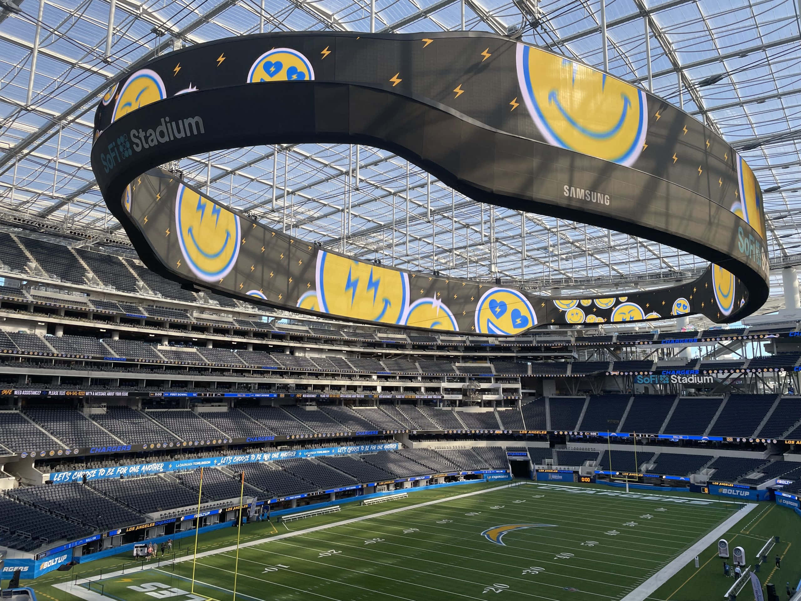 Sofi Stadium 桌布 壁紙 — 一個巨大的弧形數位螢幕顯示黃色笑臉，懸掛在玻璃屋頂下的SoFi體育場空蕩蕩的綠色足球場上方。