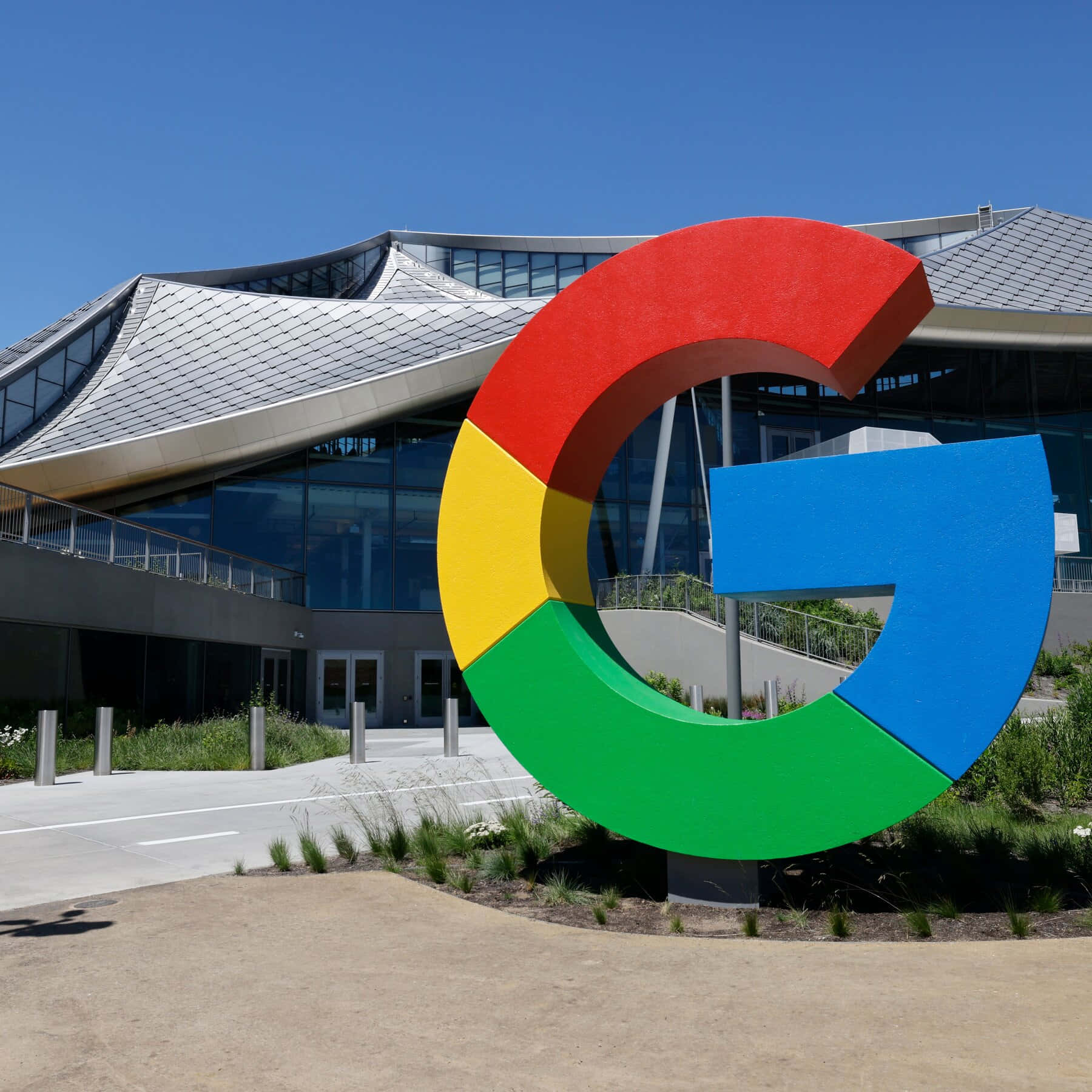 google 桌布 壁紙 — 一個巨大的彩色Google G標誌醒目地矗立在一座現代玻璃和金屬建築前，背景是清澈的藍天。