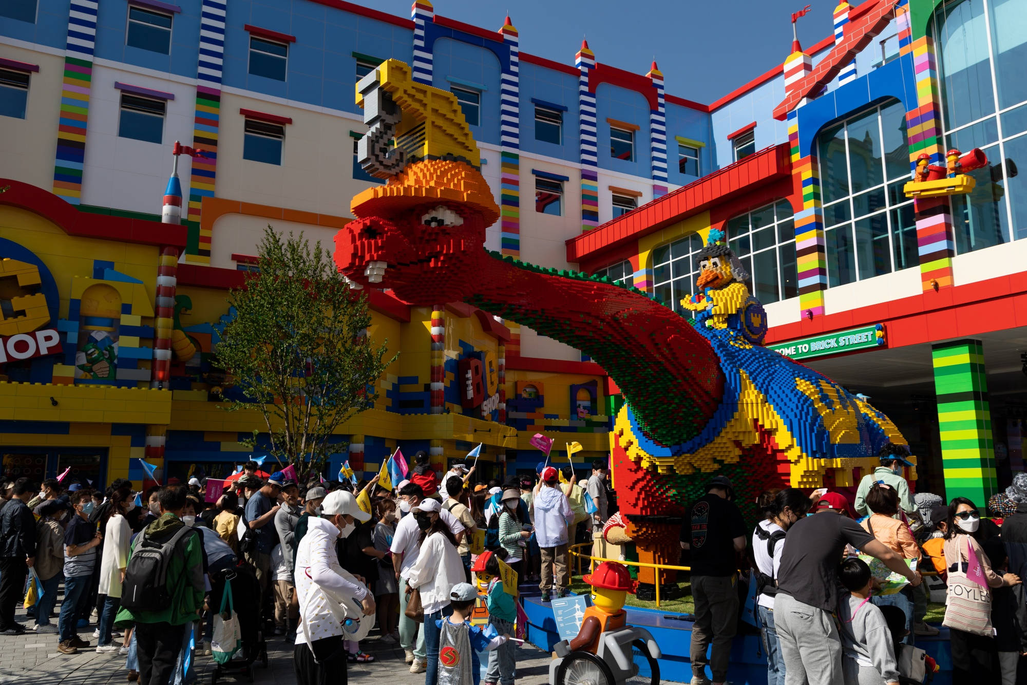 小孩動圖 桌布 壁紙 — 一個巨大的彩色LEGO恐龍雕塑在明亮的陽光下，聳立於充滿活力的LEGO主題建築前，俯瞰著人群。