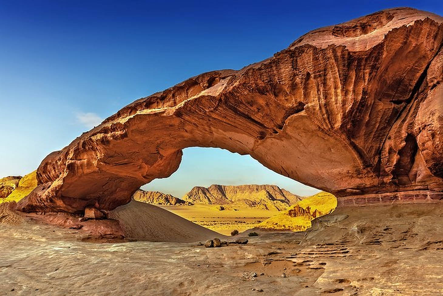 Jordan 桌布 壁紙 — 一個巨大的微紅色砂岩拱門主宰著陽光明媚的沙漠峽谷，遠處有平頂山，天空湛藍。