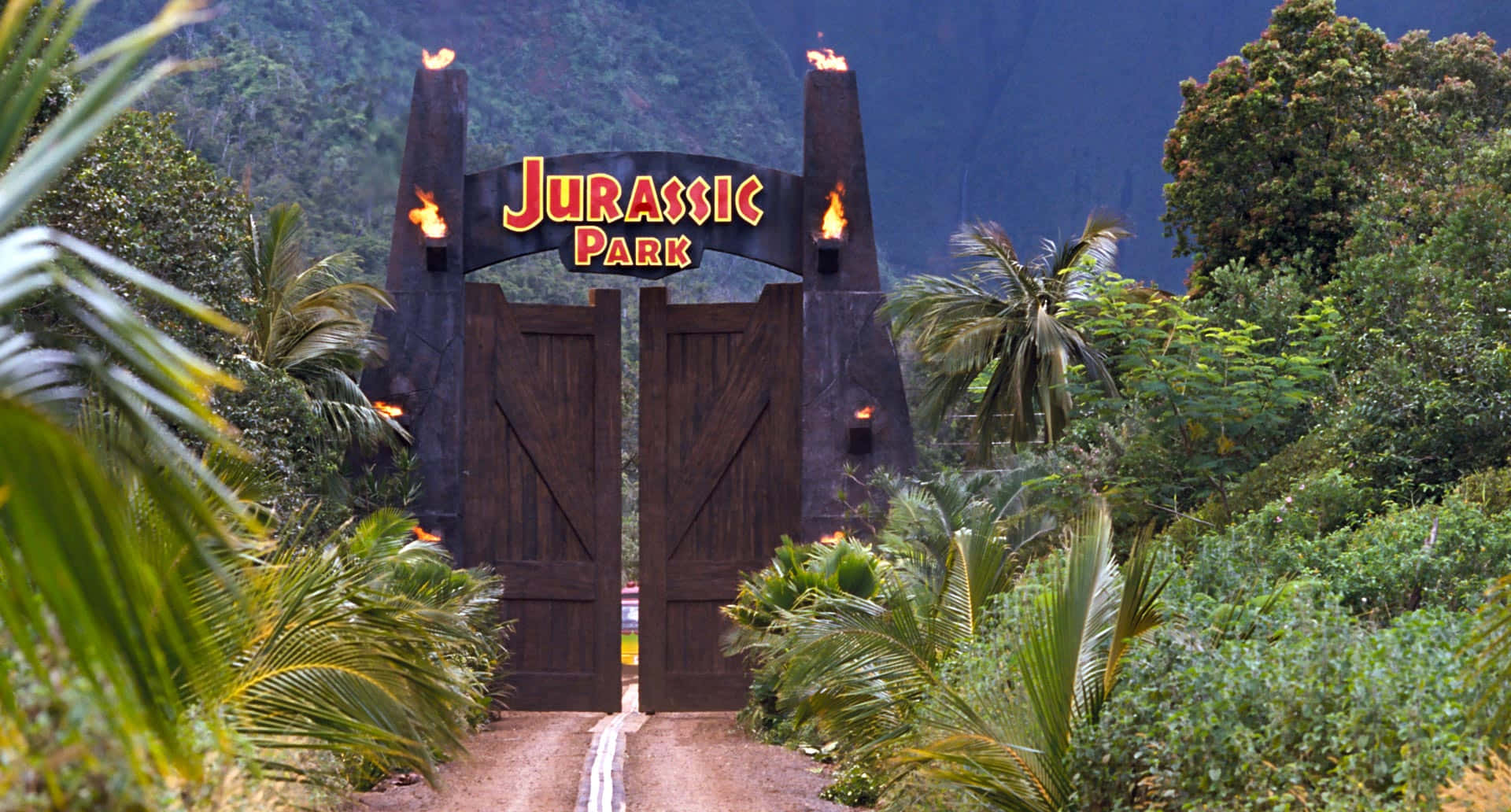 Jurassic Park Zoom 桌布 壁紙 — 一個巨大的木門，帶有火焰火炬和發光的侏羅紀公園標誌，矗立在茂密的熱帶植物中，天空陰暗。