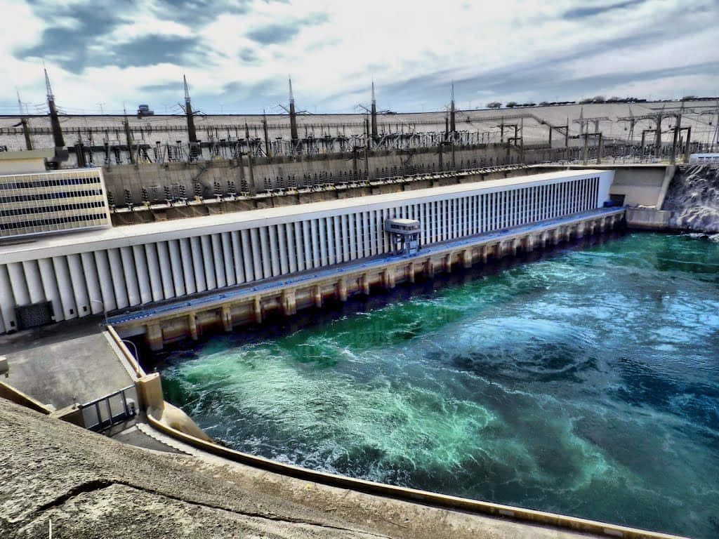 水 桌布 壁紙 — 一個巨大的混凝土水力發電水壩在陰天之下將藍綠色的水排放到湍急的河流中。