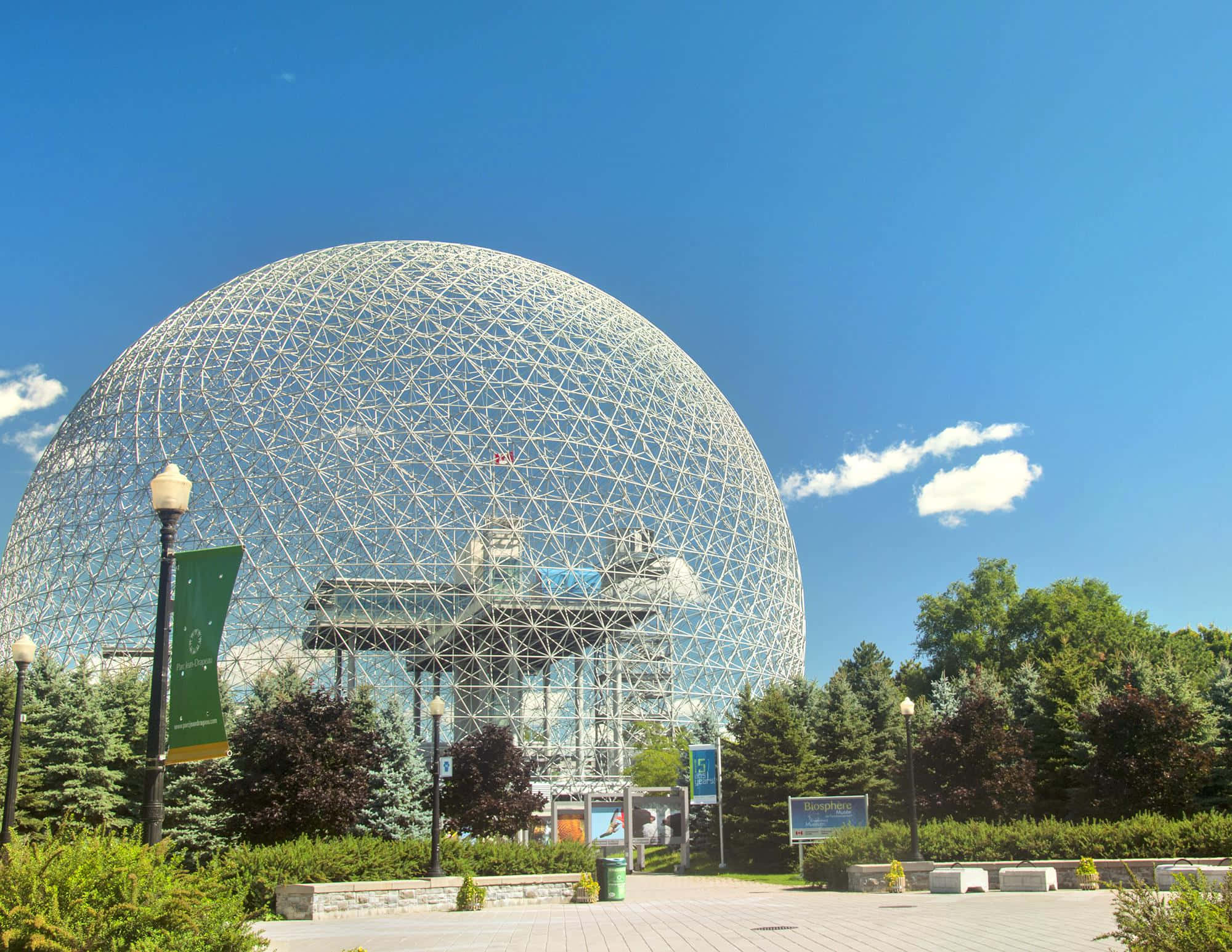 Biosphere 桌布 壁紙 — 一個巨大的測地圓頂，帶有白色網格結構，矗立在晴朗的藍天下，周圍環繞著綠樹和鋪砌的廣場。