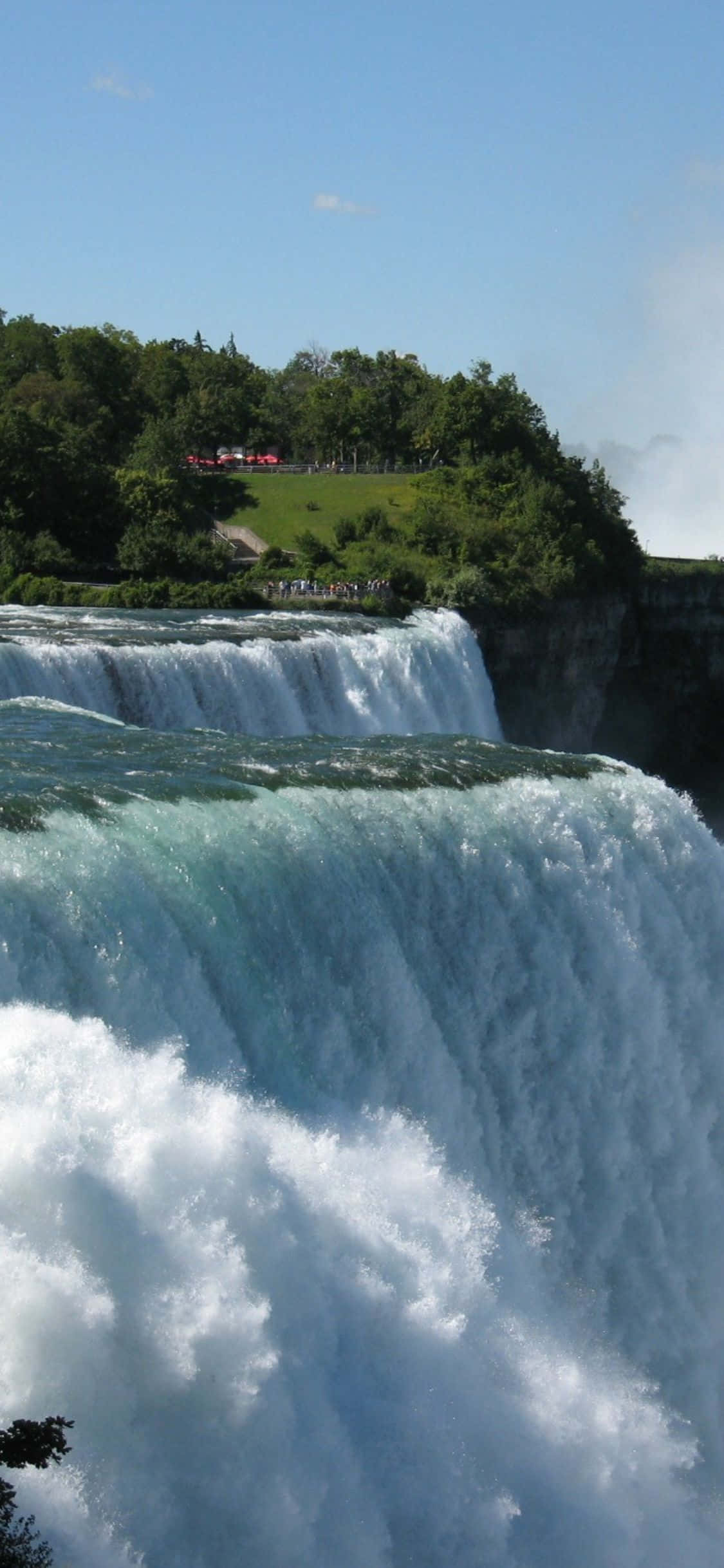 Niagara Falls 桌布 壁紙 — 一個巨大的瀑布從懸崖上強力傾瀉而下，周圍綠意盎然，上方是清澈的藍天。