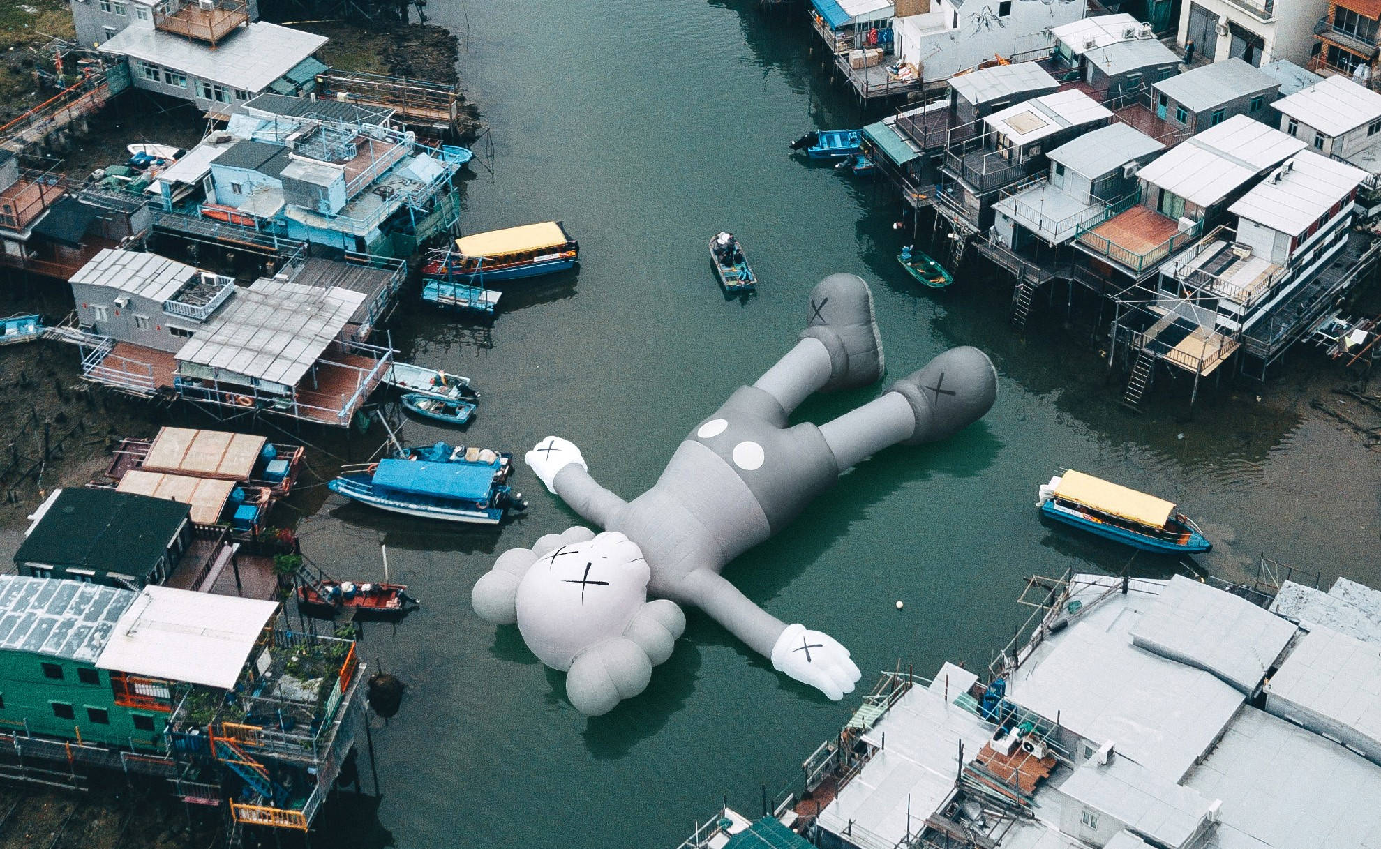 KAWS 桌布 壁紙 — 一個巨大的灰色KAWS充氣公仔在運河中平靜地漂浮，周圍是高腳屋和小船。