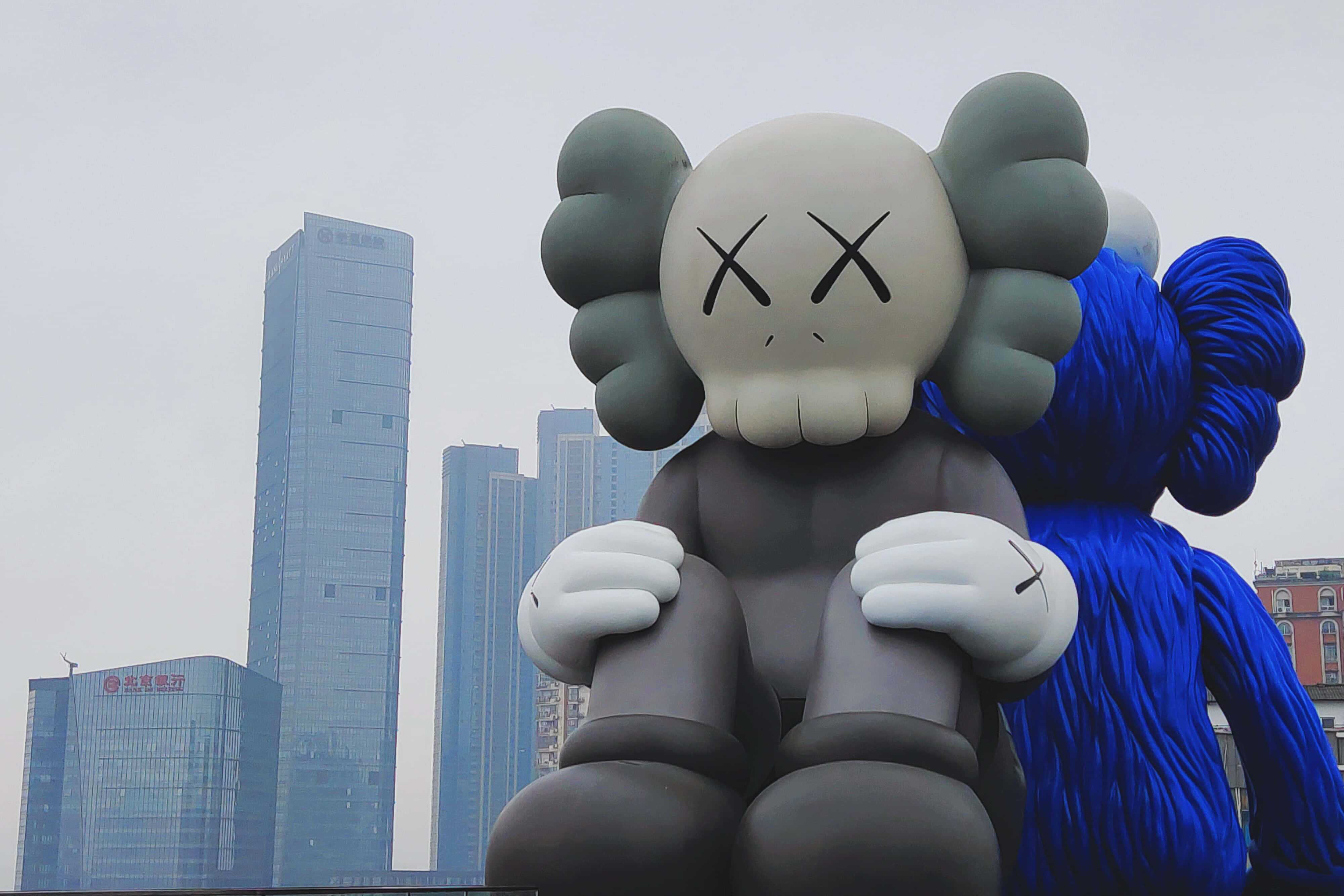 KAWS 桌布 壁紙 — 一個巨大的灰色KAWS雕塑，帶有X形眼睛，在陰天背景下，現代玻璃摩天大樓前靜靜地坐著。