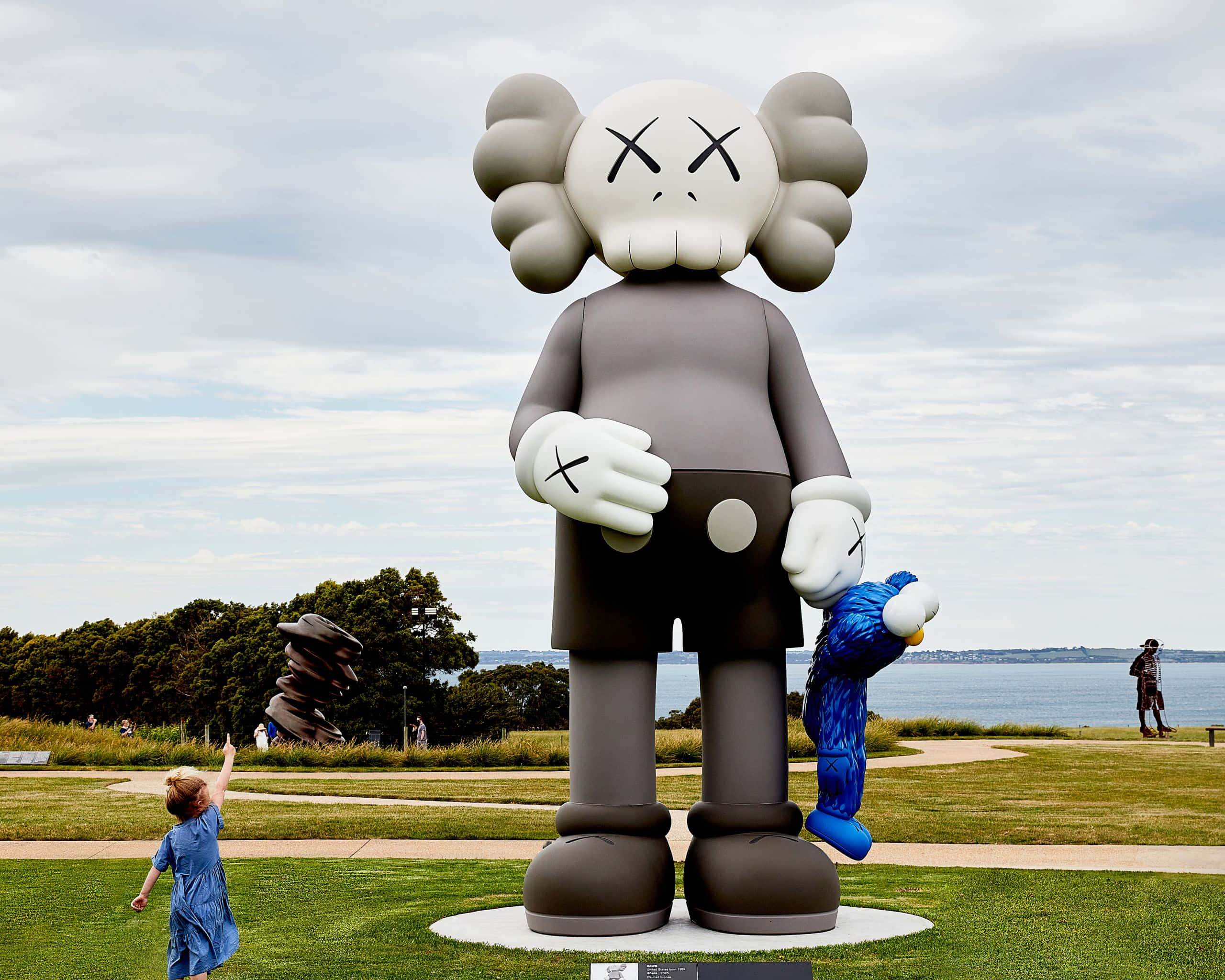 KAWS 桌布 壁紙 — 一個巨大的灰色KAWS雕塑，帶有X形眼睛，站在水邊的草地上，一個穿藍色衣服的孩子正伸手觸碰它。