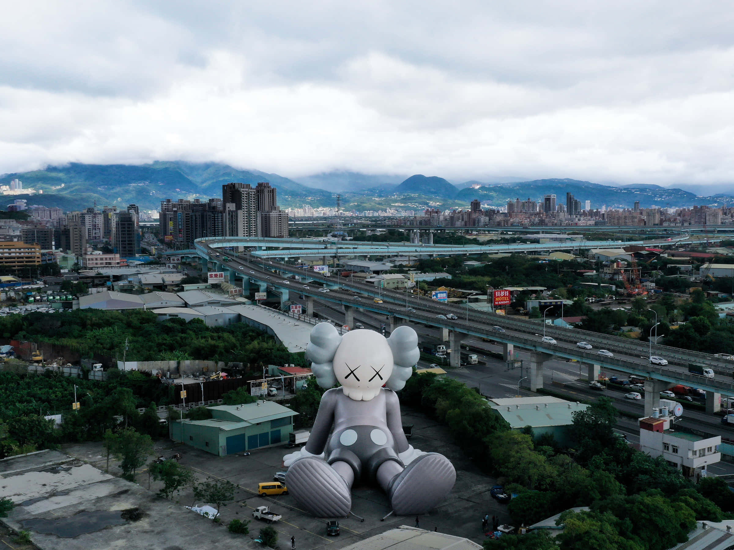 Kaws Holiday 桌布 壁紙 — 一個巨大的灰色KAWS雕塑坐落於城市地塊上，俯瞰著擁有高速公路和遠處山脈的廣闊城市，天空多雲。
