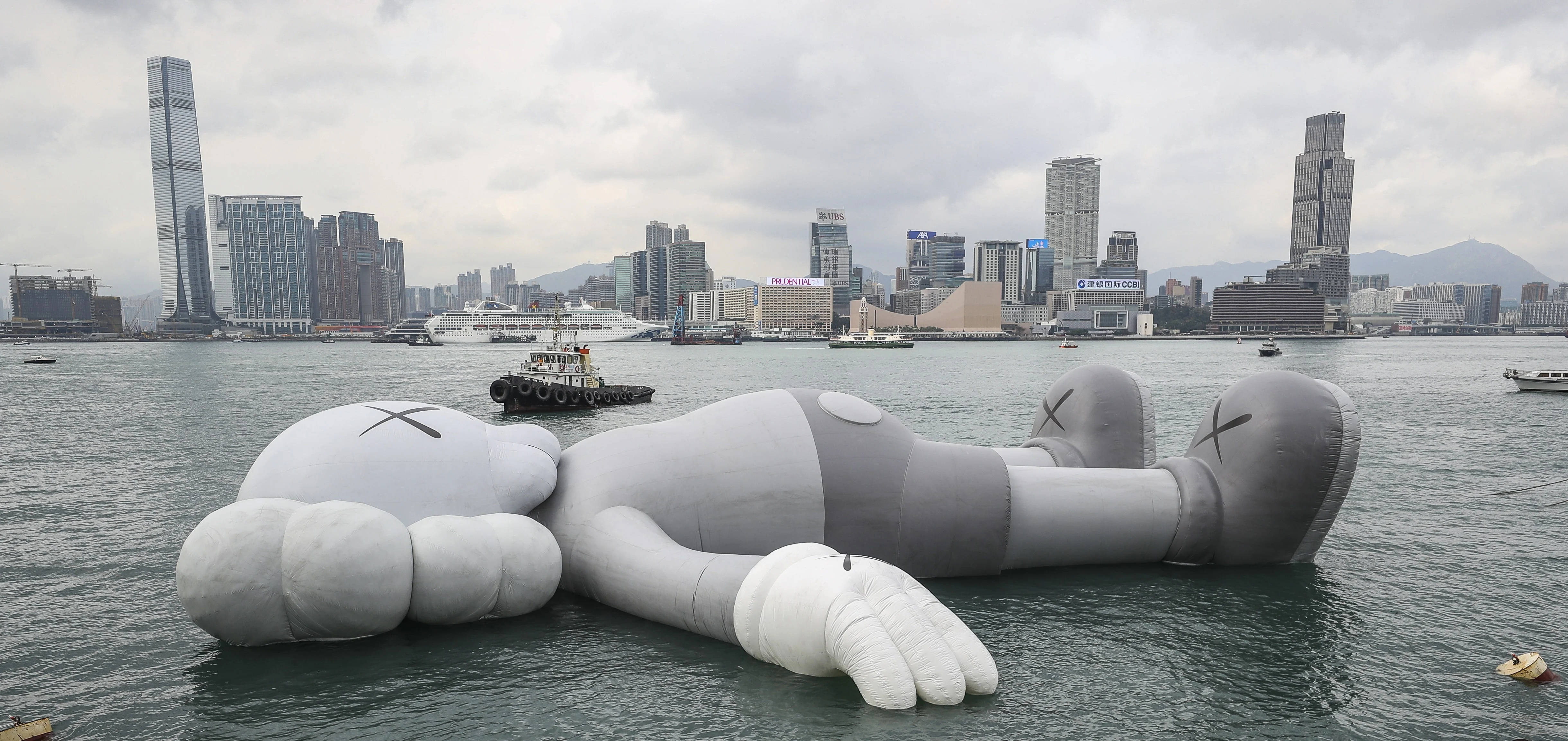KAWS 桌布 壁紙 — 一個巨大的灰色充氣KAWS雕塑在陰天背景下，在港口水中寧靜地漂浮，背後是密集的現代城市天際線。