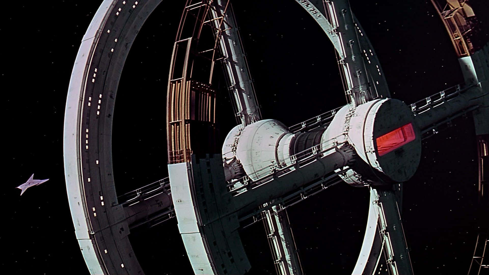 2001 A Space Odyssey 桌布 壁紙 — 一個巨大的環形太空站，帶有複雜的金屬結構和發光的紅燈，漂浮在佈滿星星的虛空中，一艘小型太空船在附近滑行。