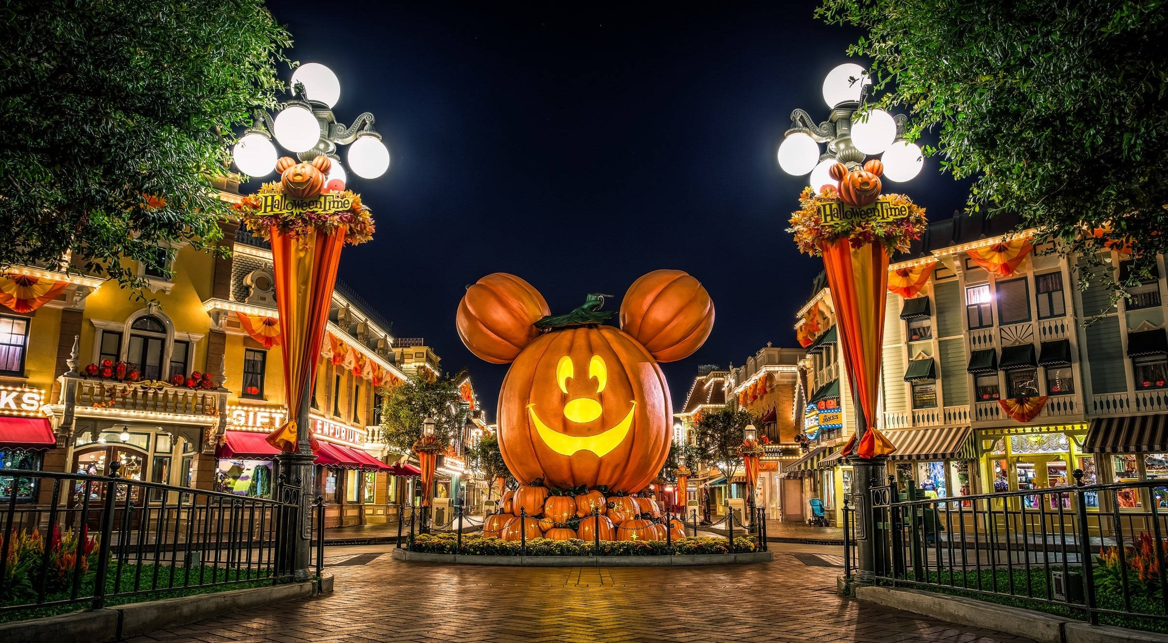 Disneyland Desktop 桌布 壁紙 — 一個巨大的發光米奇老鼠南瓜中心裝飾品，照亮了夜晚裝飾著燈柱和建築物的節日萬聖節街道。