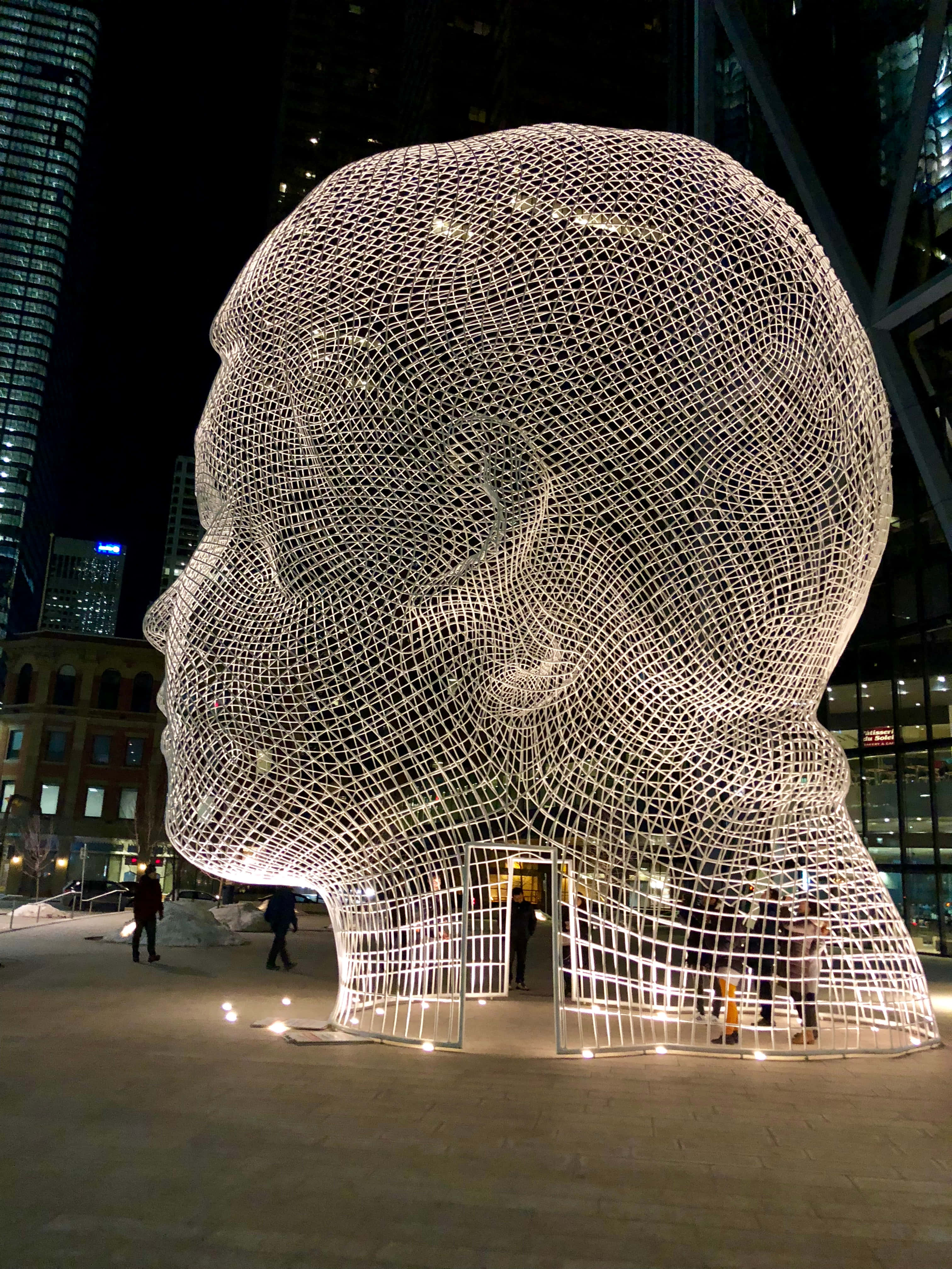 Installation Art 桌布 壁紙 — 一個巨大的發光線框頭部雕塑在夜晚的雪城市廣場中發光，周圍環繞著摩天大樓。