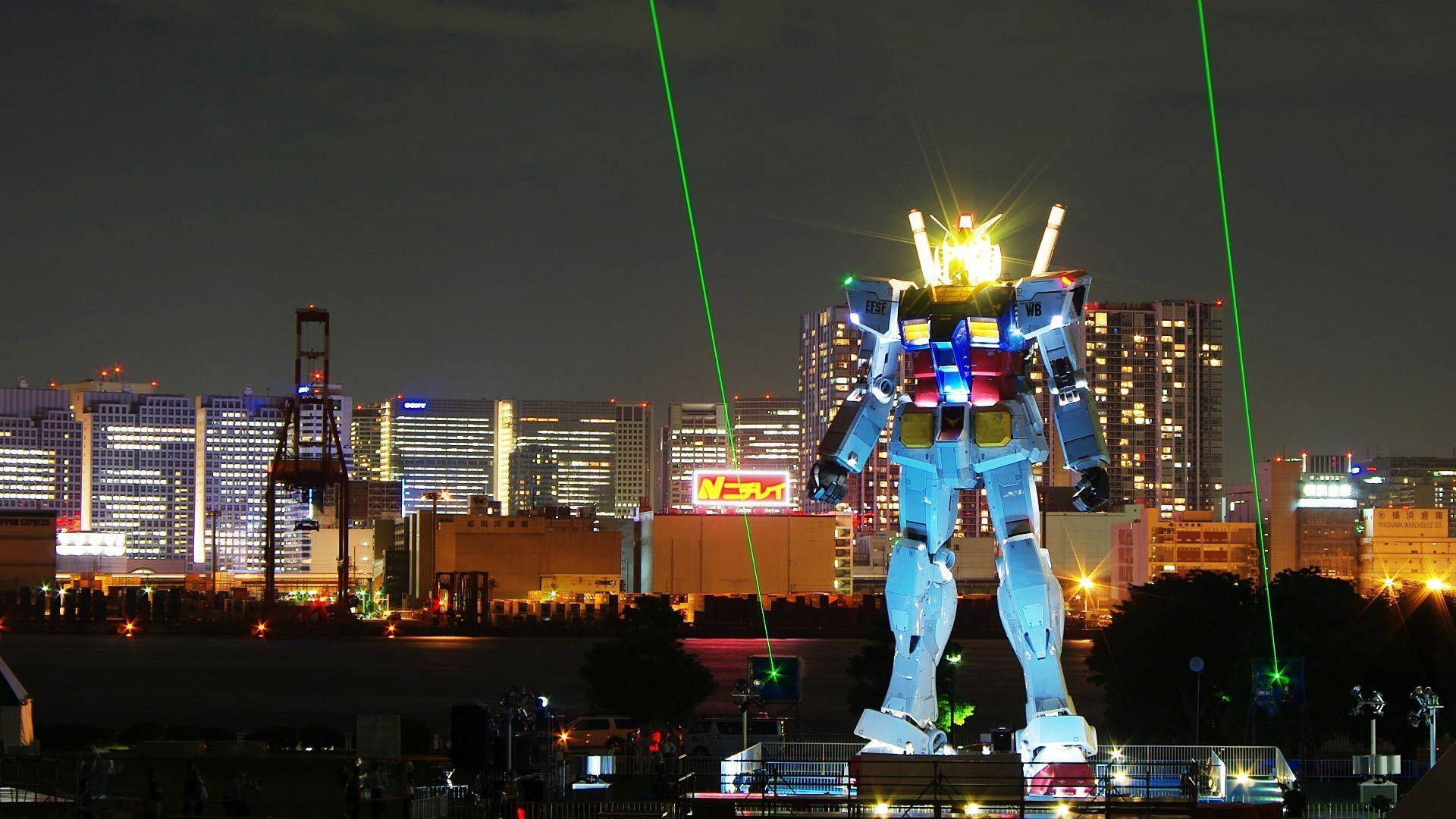 東京 桌布 壁紙 — 一個巨大的發光藍色鋼彈雕像在夜間亮起，從其肩膀射出綠色雷射光束，映襯著城市天際線。