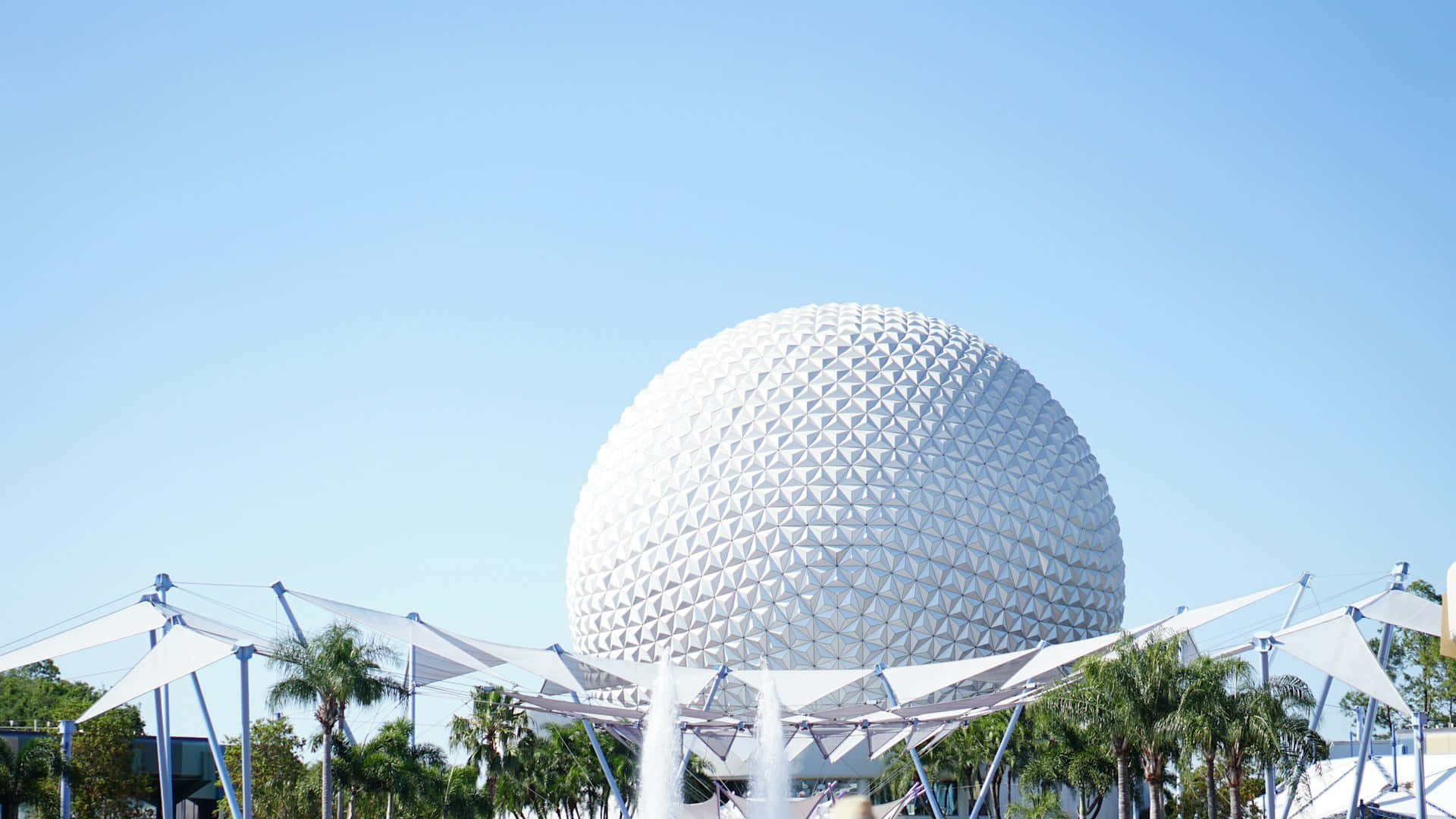 Orlando 桌布 壁紙 — 一個巨大的白色測地線球體主宰著明亮的藍天，周圍環繞著棕櫚樹和現代白色遮陽篷，下方有噴泉。