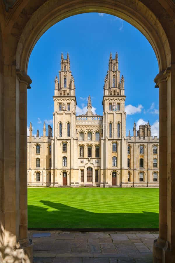 England 桌布 壁紙 — 一個巨大的石拱門框住了一座宏偉的哥德式大學建築，雙塔尖頂襯著明亮的藍天，下方是充滿活力的綠色草坪。