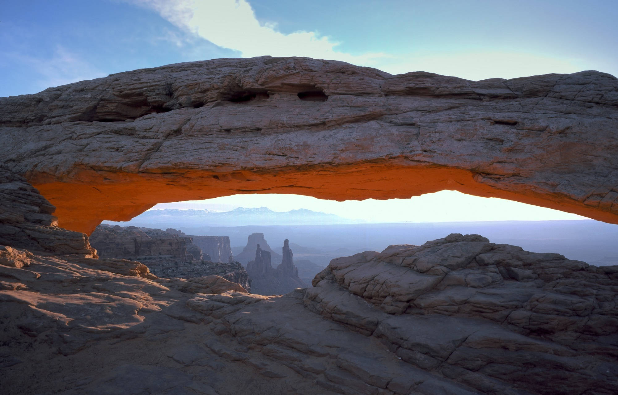 Canyonlands National Park 桌布 壁紙 — 一個巨大的砂岩拱門在日出時發出橙色光芒，框住遠處的平頂山，在清澈的藍天下。
