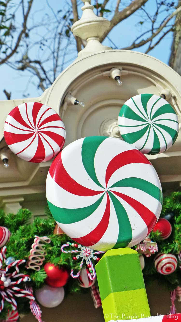 Disney Christmas iPad 桌布 壁紙 — 一個巨大的糖果杖米奇老鼠頭，帶有紅色、白色和綠色的漩渦，矗立在一個裝飾著花環和裝飾品的節日拱門前。
