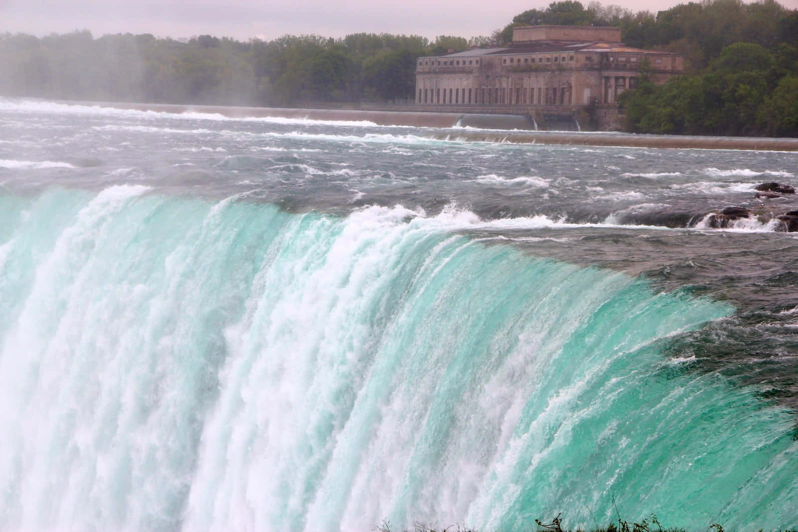 Niagara Falls 桌布 壁紙 — 一個巨大的藍綠色瀑布傾瀉而下，水霧升起，一座石頭建築矗立在樹後的河岸上。