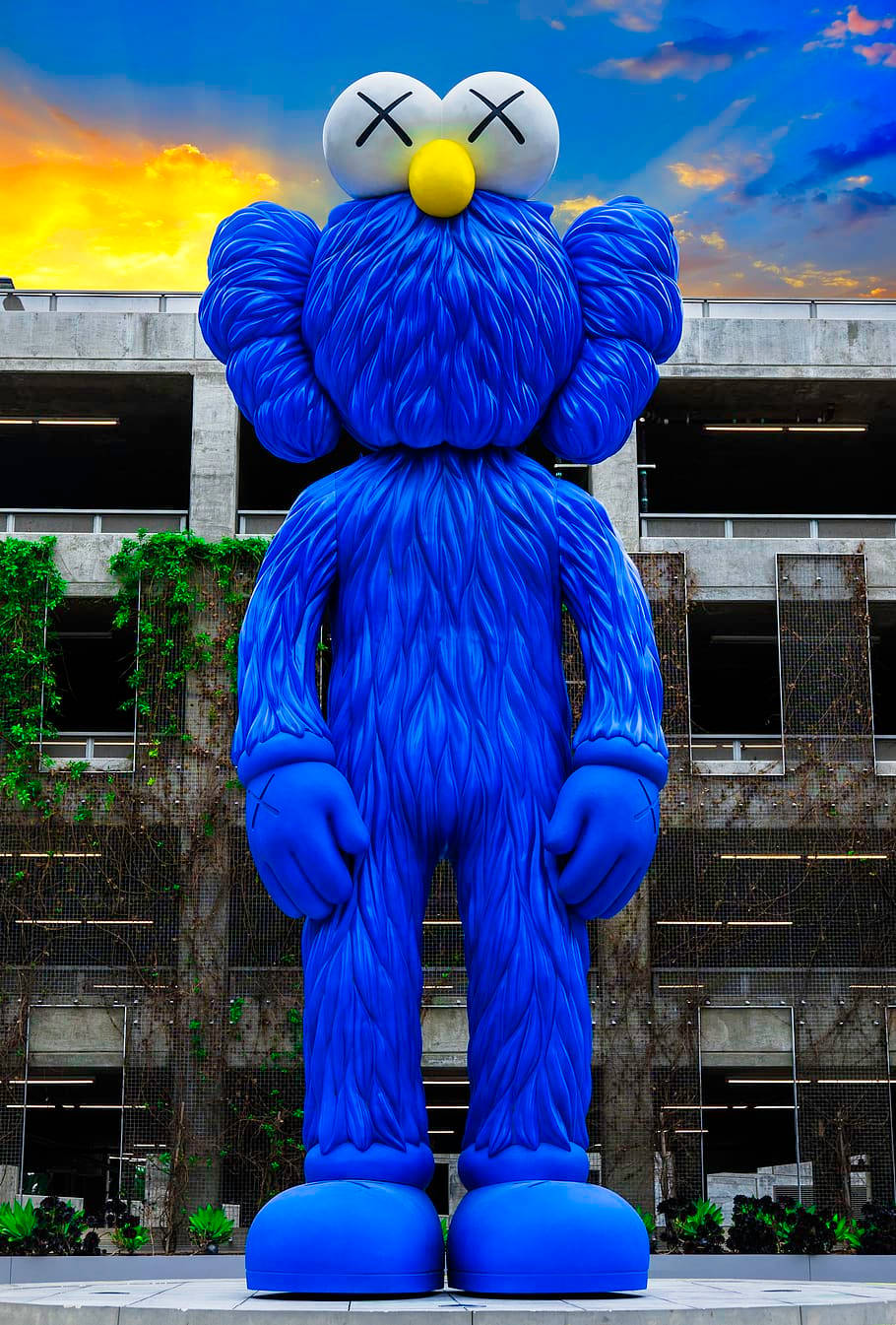 kaws 4K 桌布 壁紙 — 一個巨大的藍色KAWS雕塑，帶有X形眼睛，矗立在日落時分的混凝土停車場前。