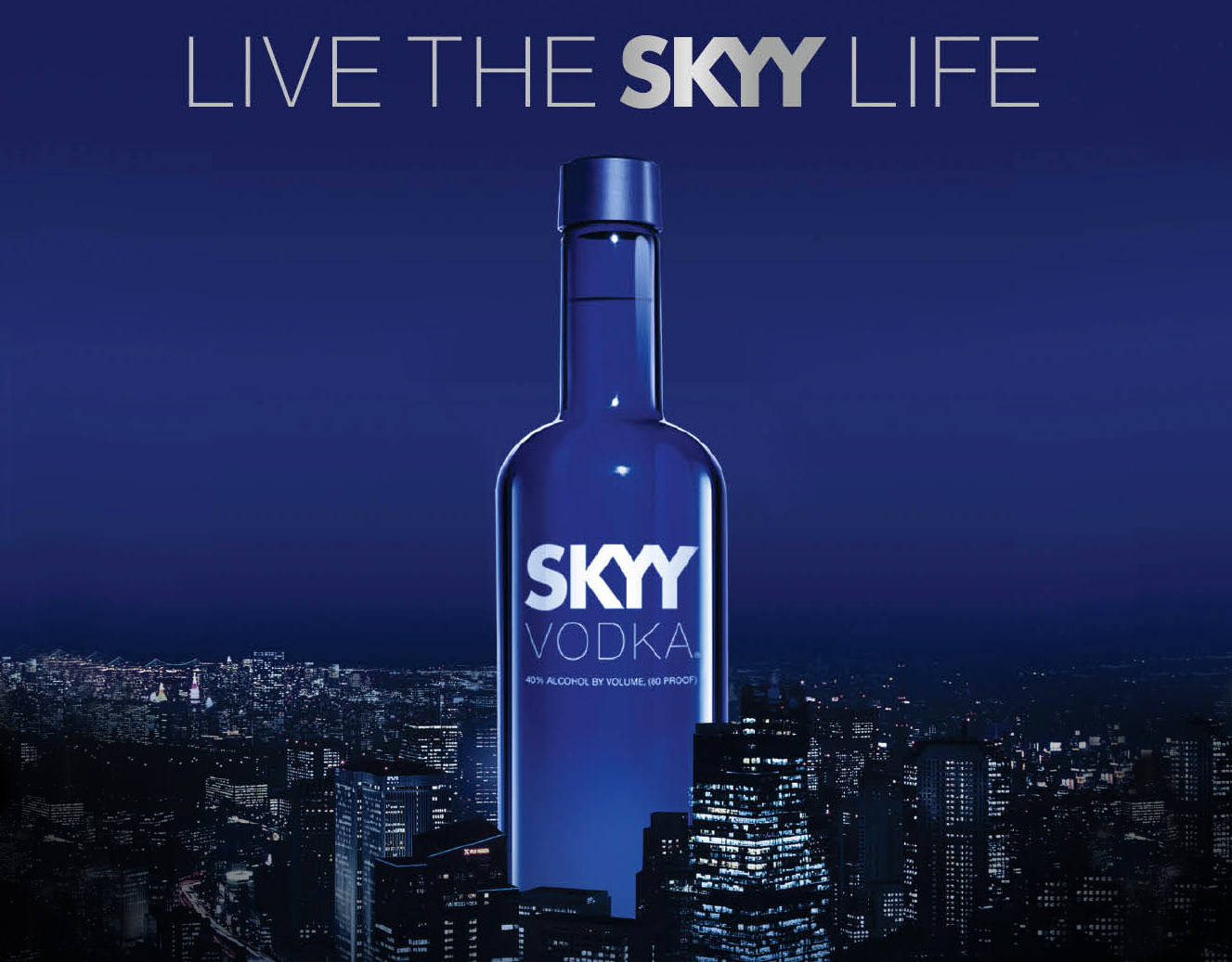 Drink 桌布 壁紙 — 一個巨大的藍色Skyy伏特加酒瓶聳立在深藍色天空下閃閃發光的夜景城市景觀之上。