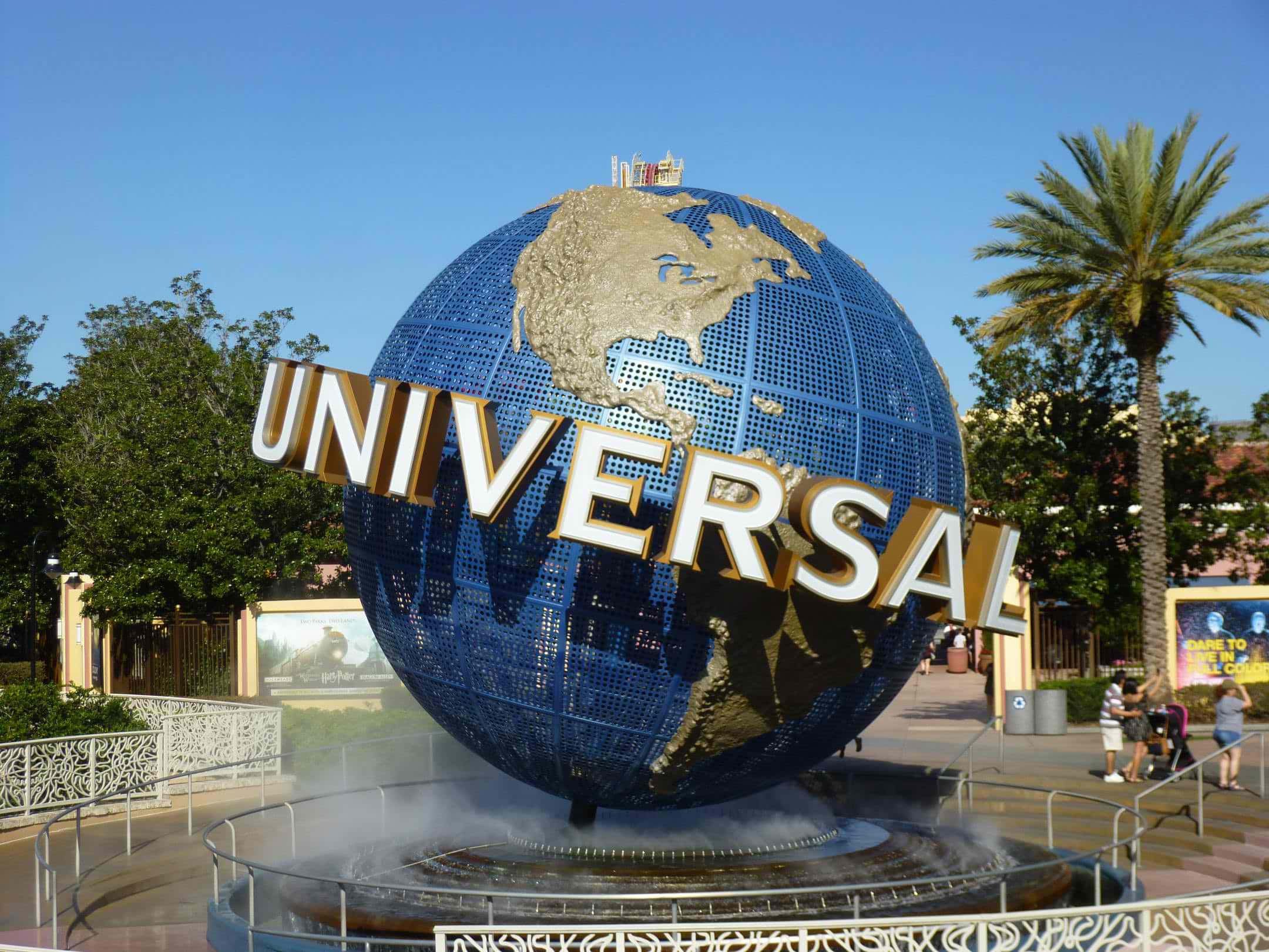 Universal 桌布 壁紙 — 一個巨大的藍色地球儀，上面有金色的大陸和「UNIVERSAL」標誌，矗立在主題公園入口處，藍天白雲，附近有棕櫚樹和遊客。