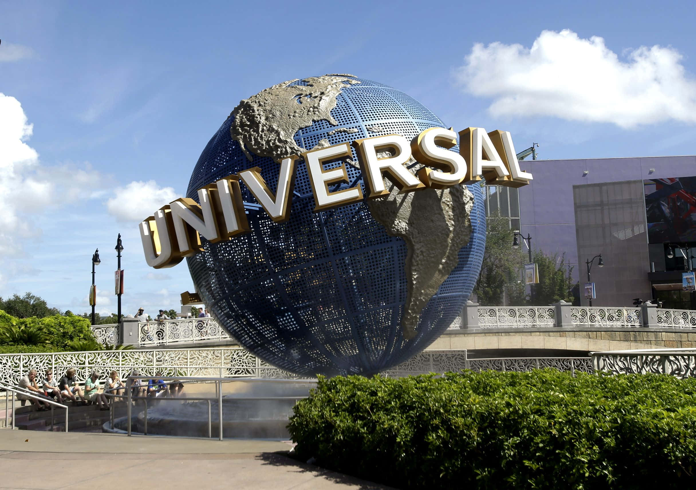 Universal 桌布 壁紙 — 一個巨大的藍色地球儀，上面有金色的「UNIVERSAL」字樣，主宰著陽光明媚的主題公園入口，下方有茂密的綠植和遊客。
