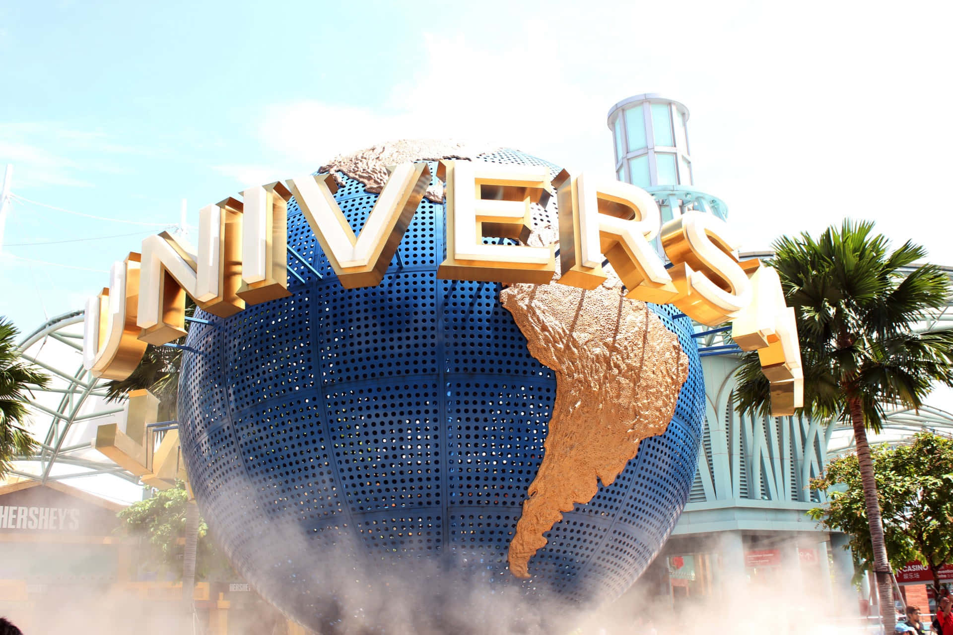 Universal 桌布 壁紙 — 一個巨大的藍色穿孔地球儀，帶有金色大陸和「UNIVERSAL」字樣，拱形橫跨在霧氣瀰漫的主題公園入口上方，兩旁種有棕櫚樹。