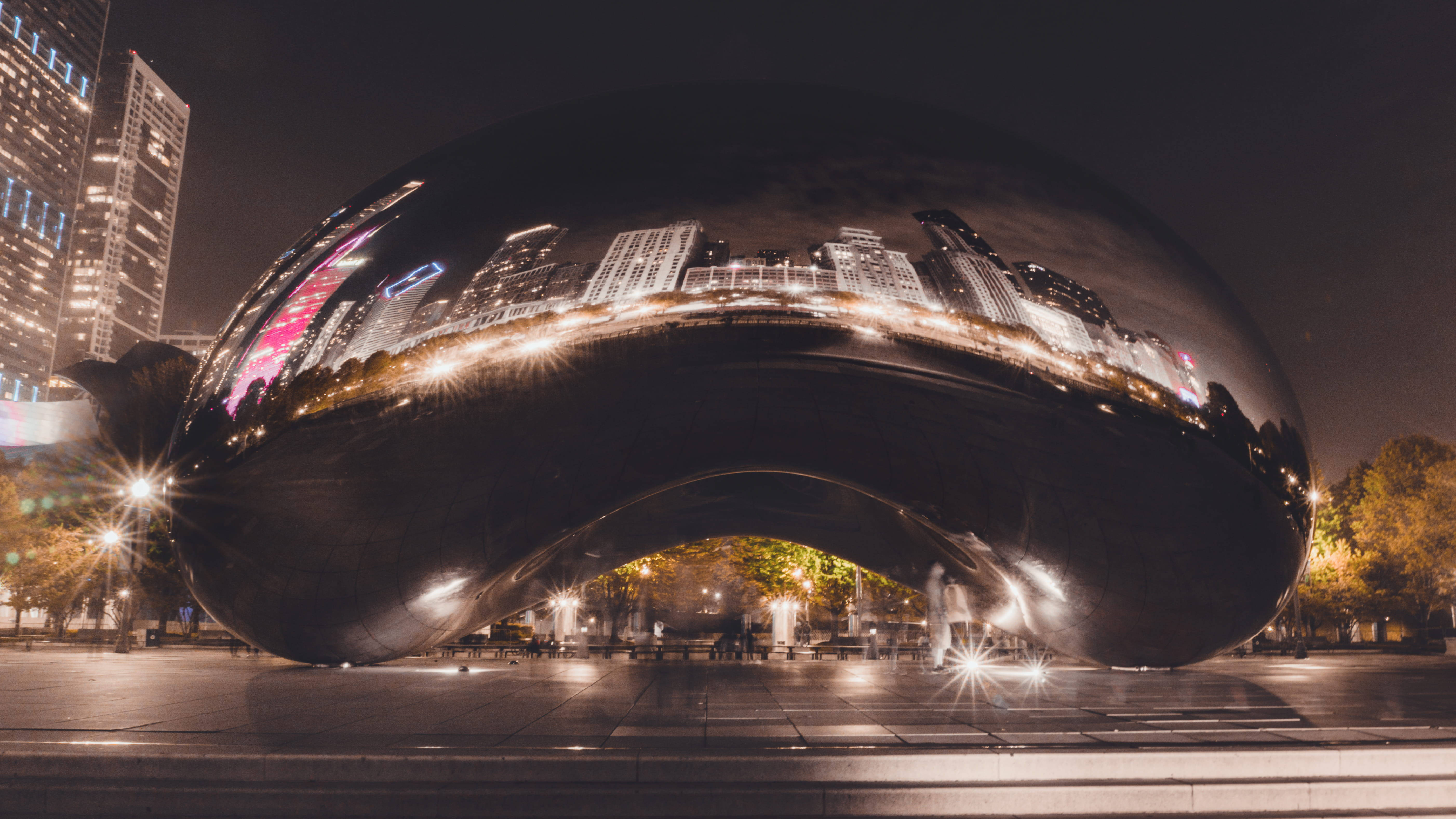 City At Night 桌布 壁紙 — 一個巨大的豆形雕塑反射出夜間充滿活力的城市天際線，下方有發光的燈光和模糊的行人。