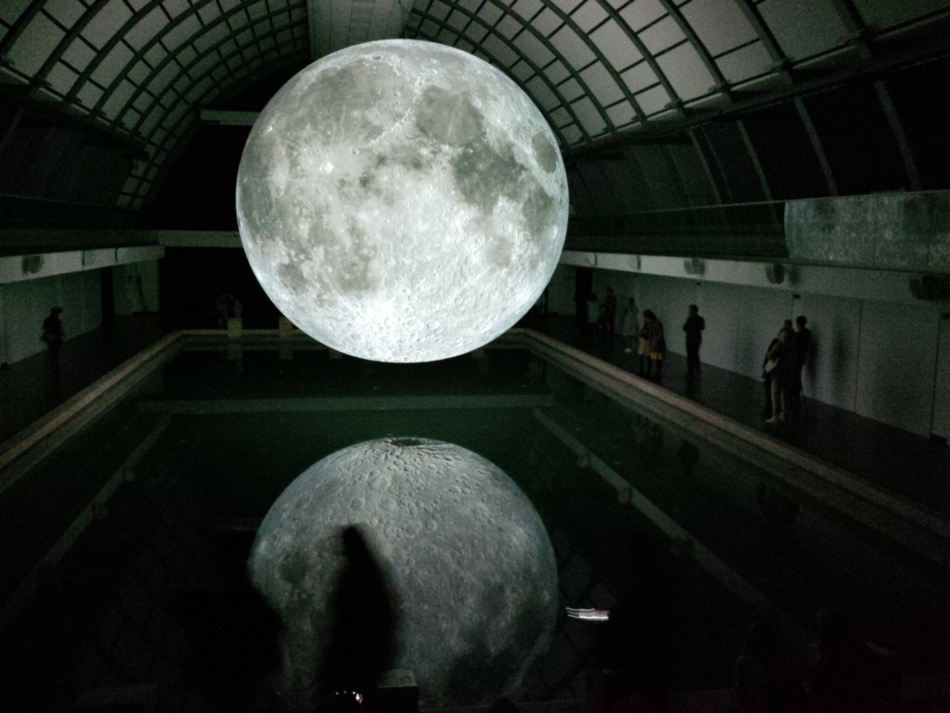 full moon 桌布 壁紙 — 一個巨大的逼真月球雕塑漂浮在黑暗工業大廳的反射池上方，有參觀者在觀察。