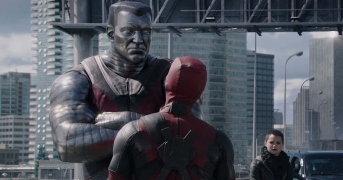 Deadpool And Colossus 桌布 壁紙 — 一個巨大的金屬人形站在一個穿著紅色套裝的人物上方，背景是摩天大樓的城市景觀和一個旁觀者。