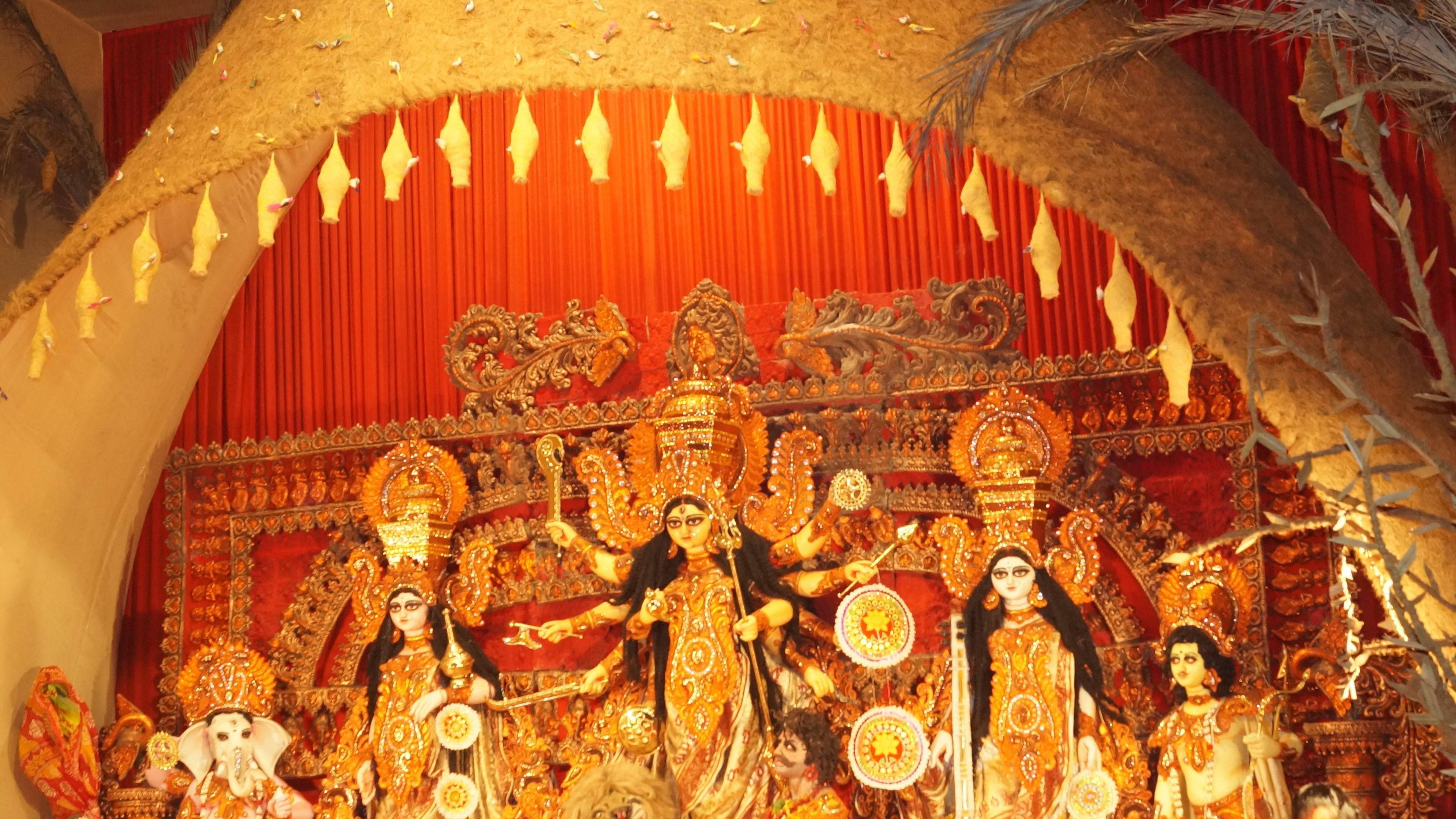 Mumbai 桌布 壁紙 — 一個巨大的金色杜爾迦普迦神像，有多隻手臂和武器，站在紅色窗簾和稻草拱門下的中央。
