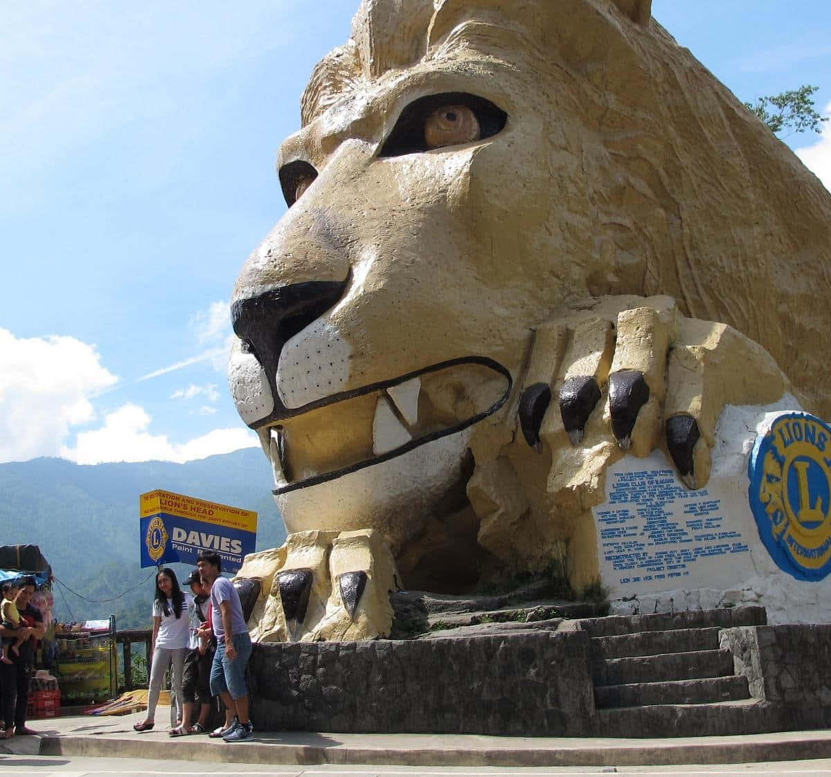 Lion Head 桌布 壁紙 — 一個巨大的金色獅頭雕塑，牙齒和爪子清晰可見，矗立在戶外，藍天白雲，背景有山巒。