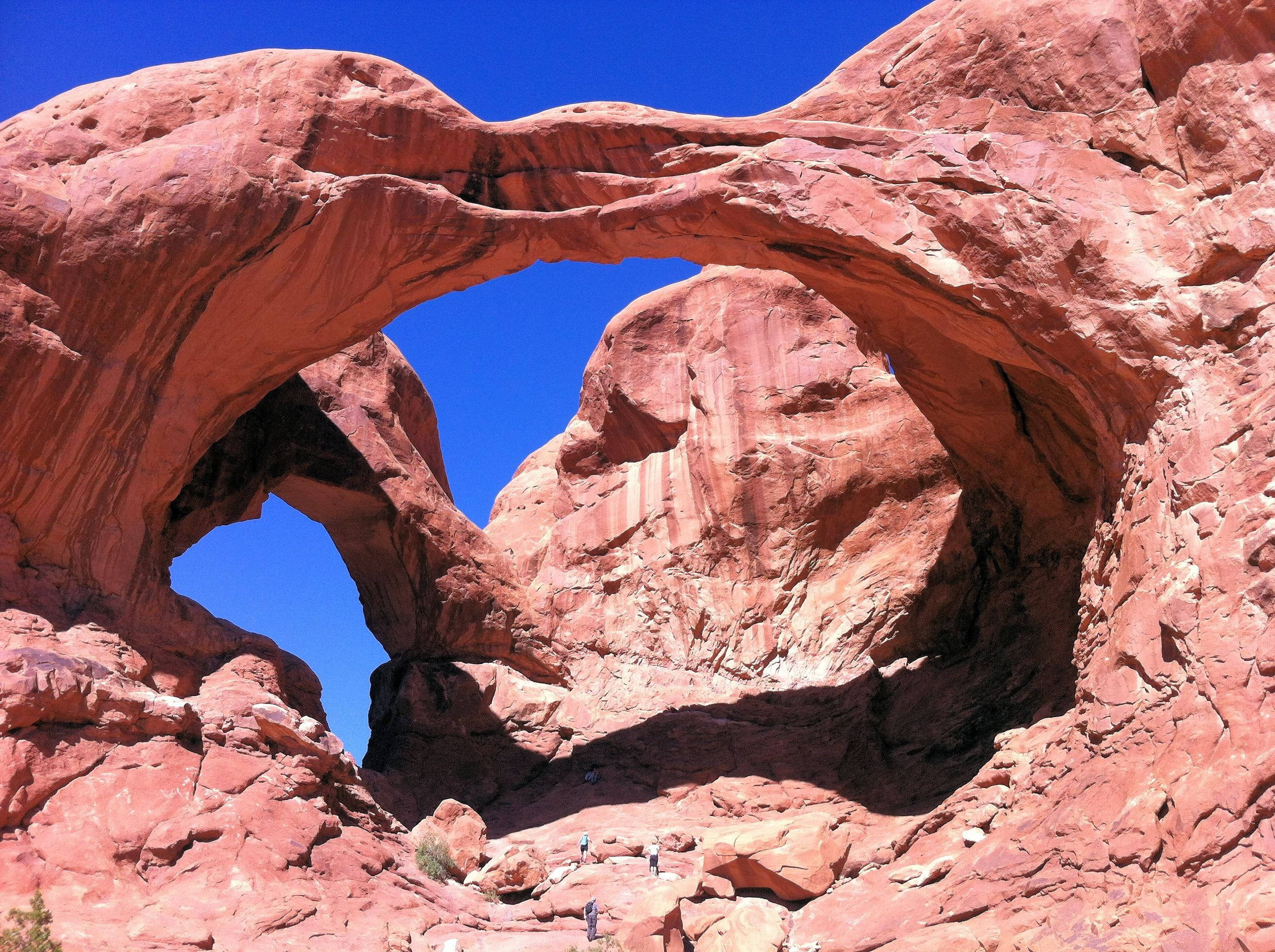 Canyonlands National Park 桌布 壁紙 — 一個巨大的雙紅石拱門框住晴朗的藍天，下方有小小的遠足者。