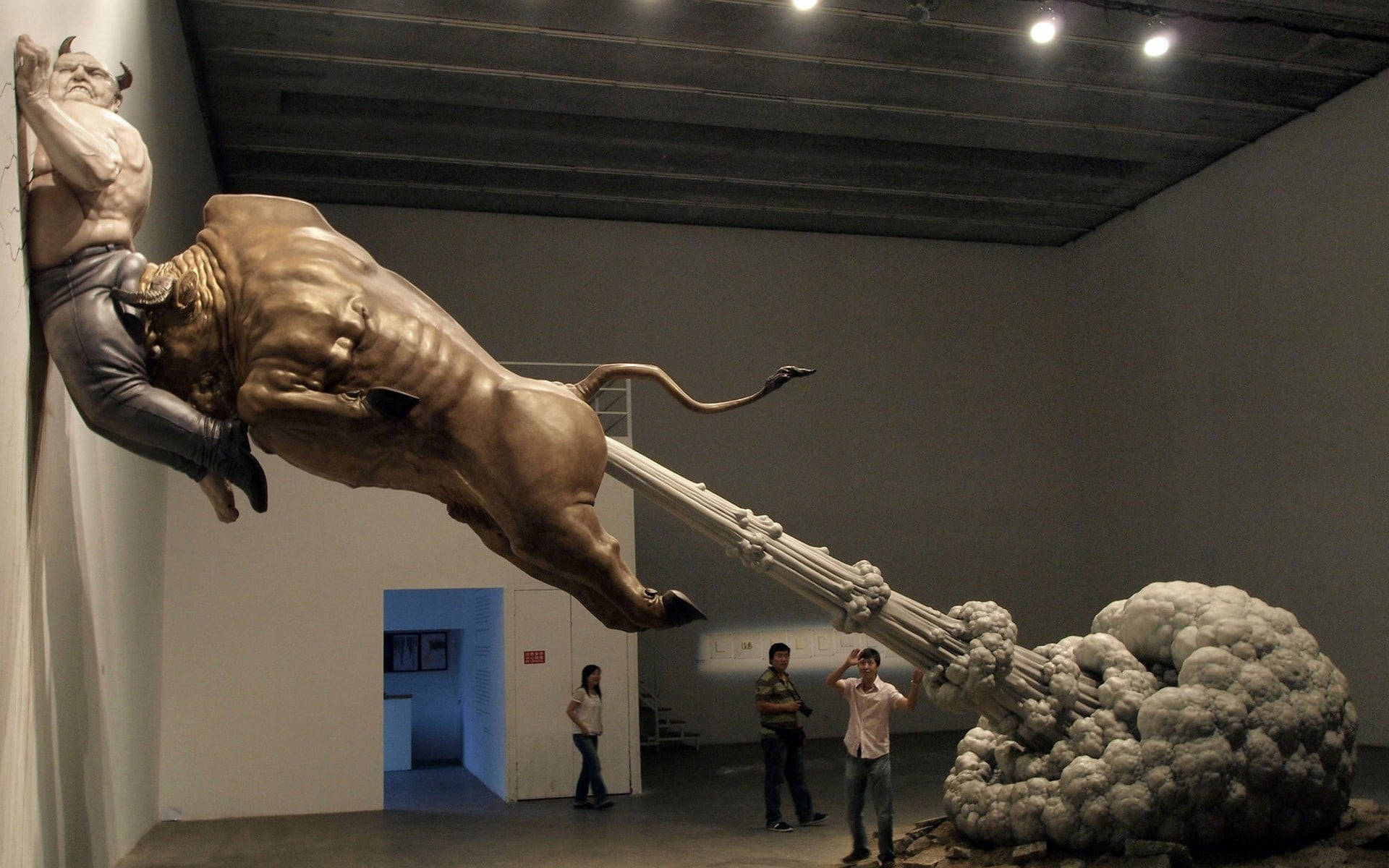 Gallery 桌布 壁紙 — 一個巨大的青銅雕塑，描繪一名男子騎著一頭衝鋒的公牛，主宰著一個現代畫廊空間，有參觀者在觀看。