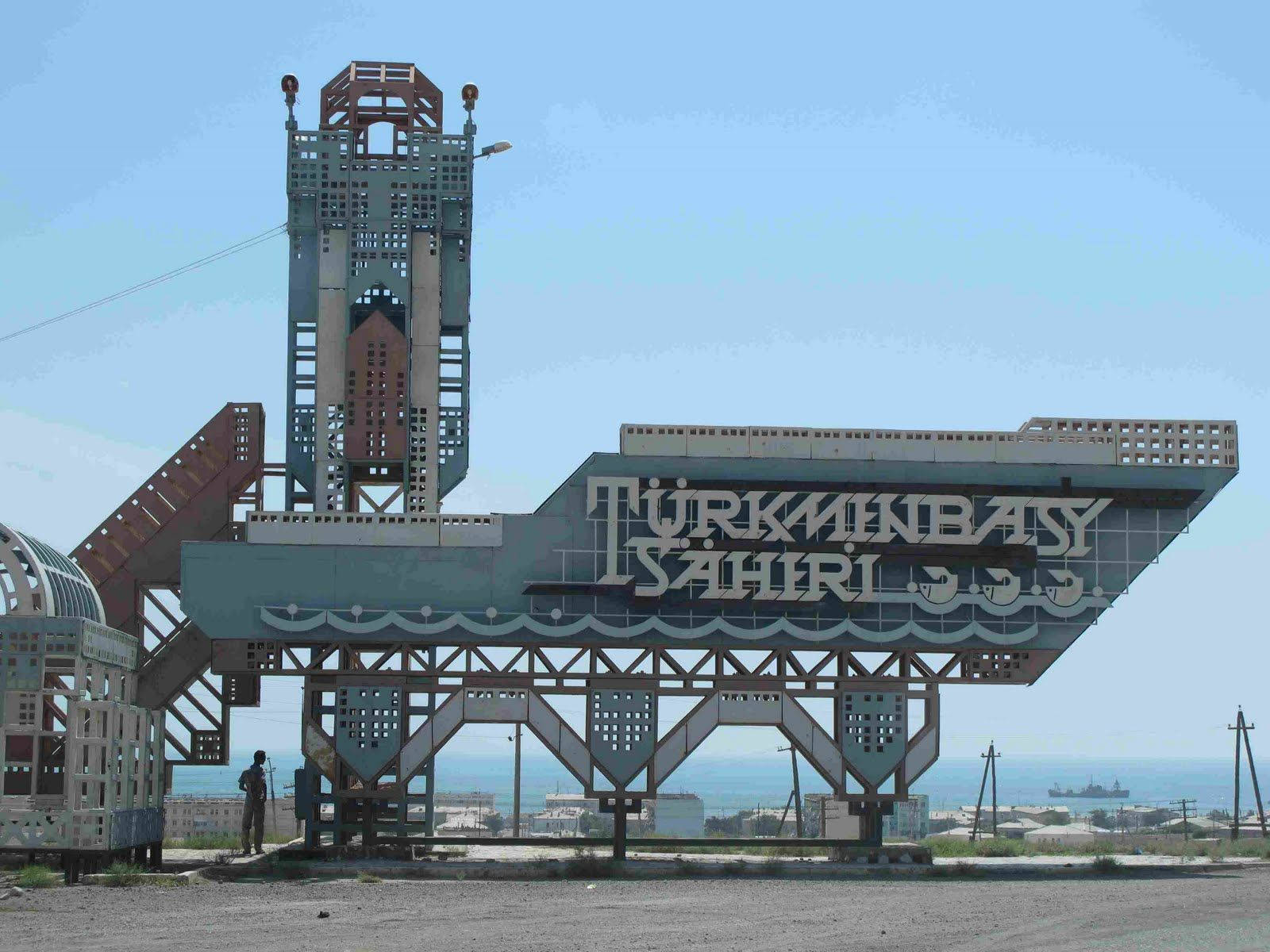 Turkmenistan 桌布 壁紙 — 一個巨大的風化標牌，上面寫著「Türkünbasy Sahiri」，在清澈的藍天、海洋和遠處船隻的背景下矗立。