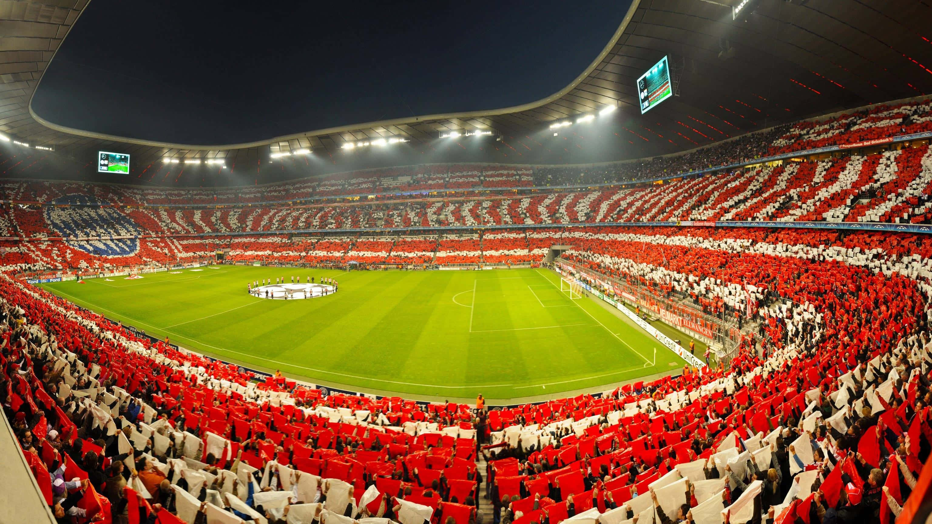 Soccer 4K 桌布 壁紙 — 一個巨大的體育場，在夜間足球比賽期間，紅白球迷在明亮的泛光燈下揮舞旗幟。