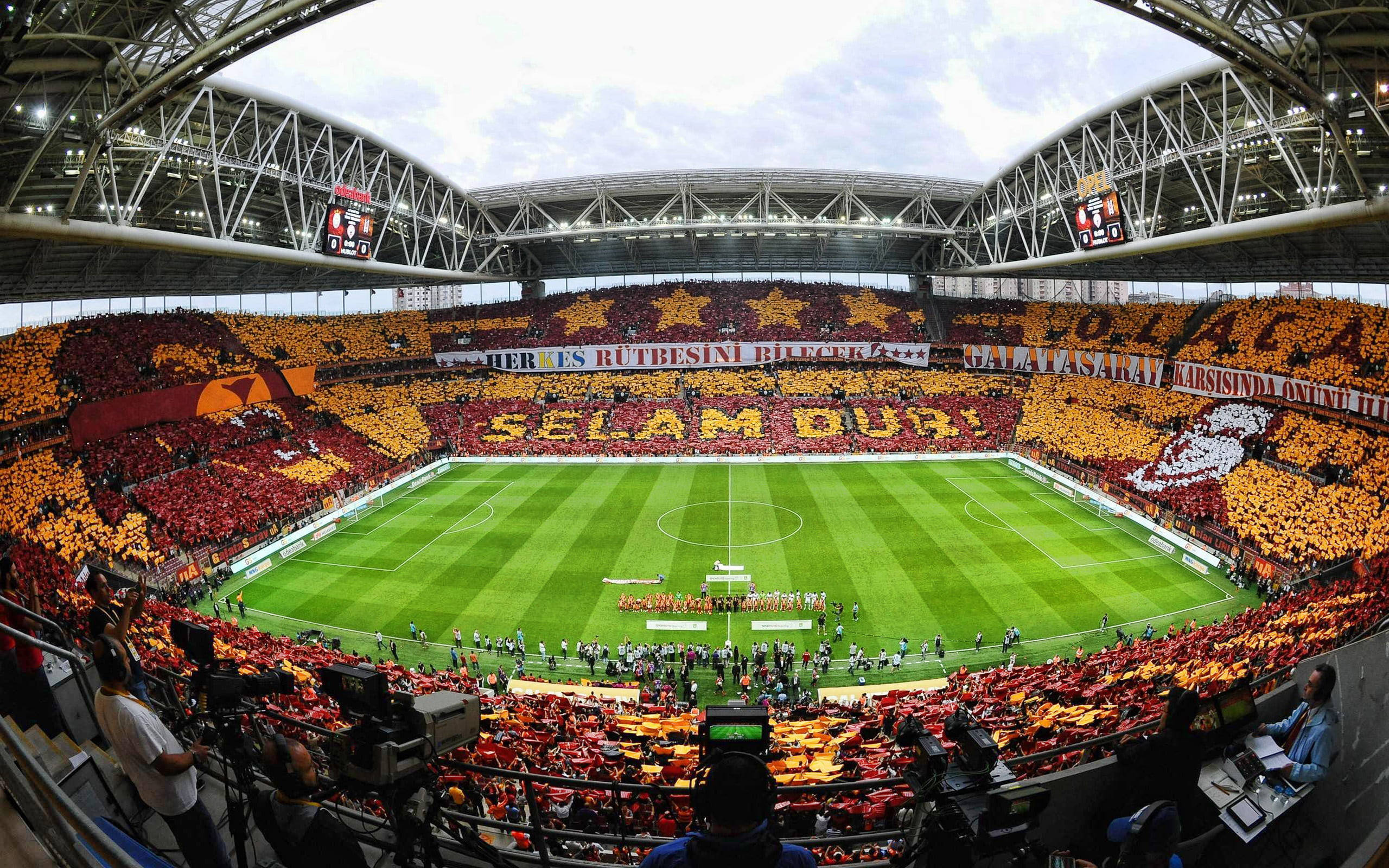 Stadium 桌布 壁紙 — 一個巨大的體育場，擠滿了身穿紅色和黃色的球迷，在充滿活力的綠色足球場上方形成巨大的字母展示。