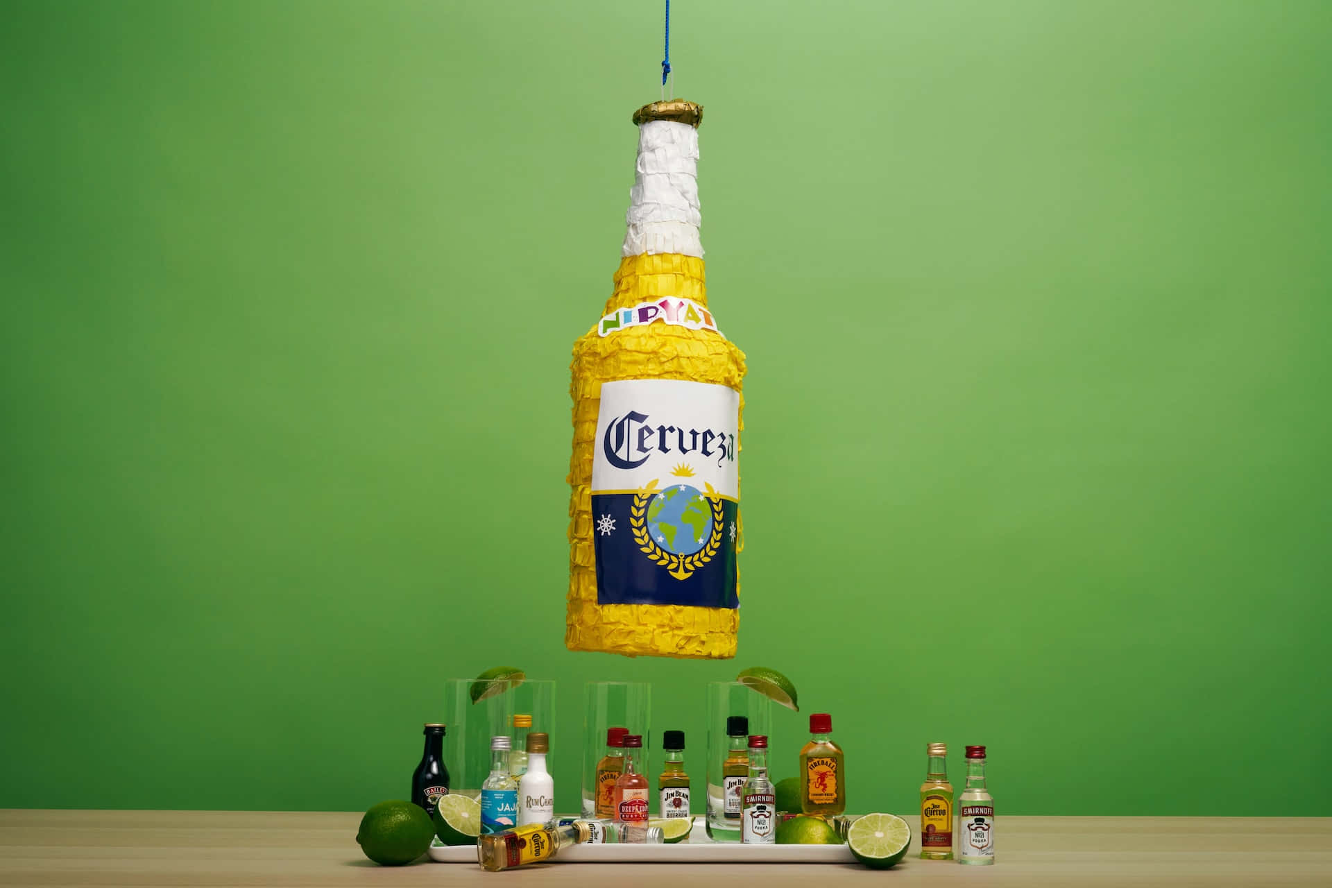 Cerveza 桌布 壁紙 — 一個巨大的黃色啤酒瓶皮納塔懸掛在吧檯上方，吧檯上有萊姆角和迷你酒瓶，放在木桌上，背景是純綠色。