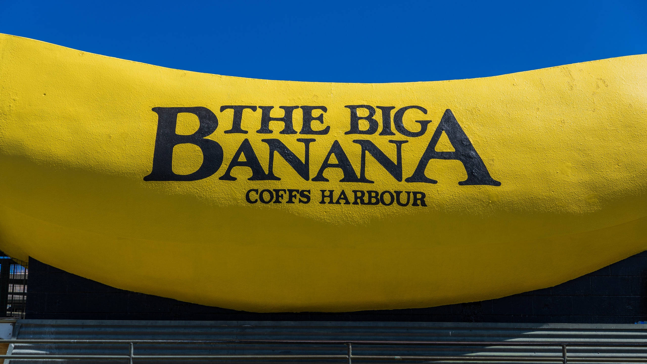 Banana 桌布 壁紙 — 一個巨大的黃色香蕉雕塑，上面有粗體黑色文字寫著「THE BIG BANANA COFFS HARBOUR」，背景是清澈的藍天。