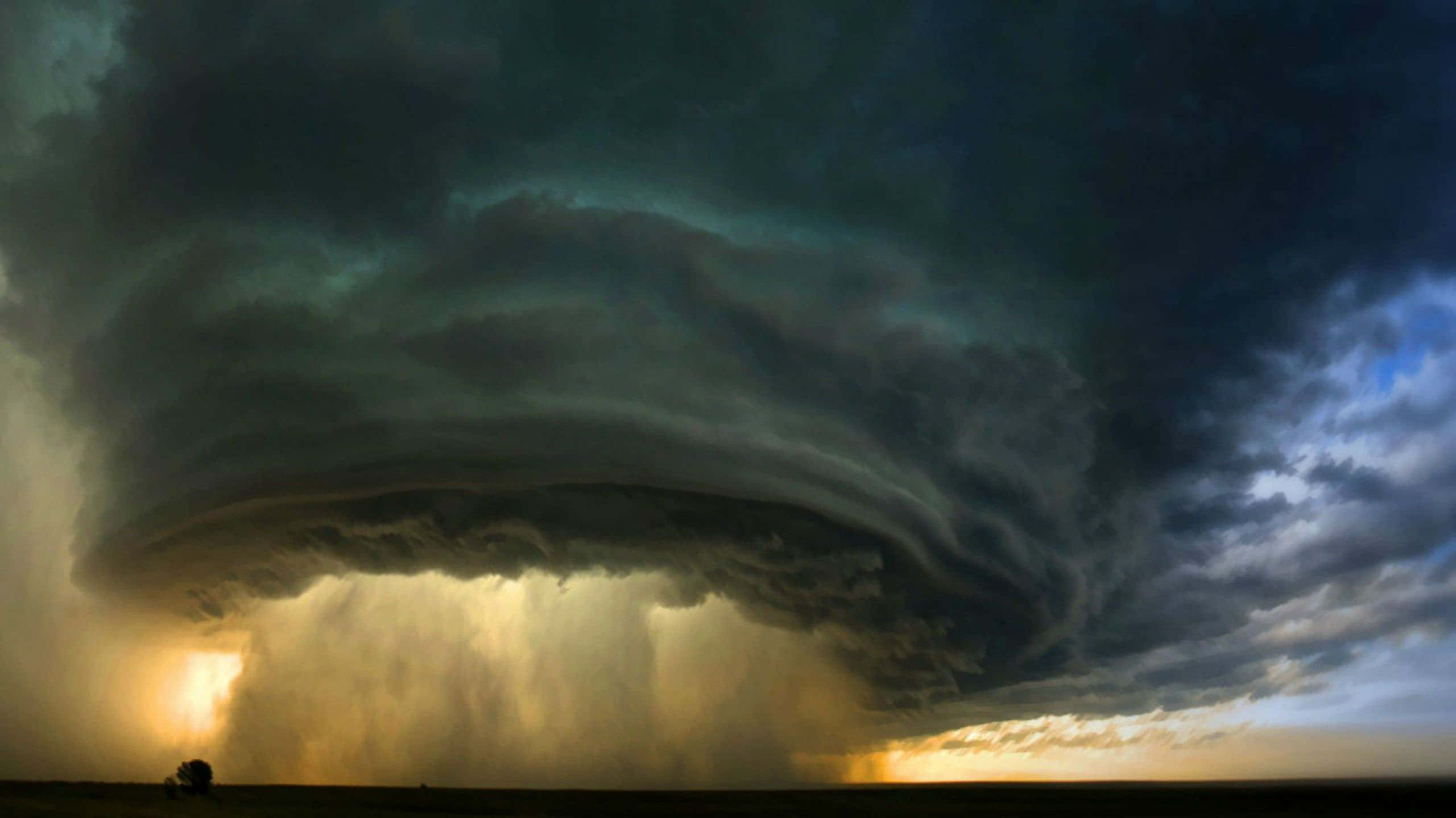 Natural Disaster 桌布 壁紙 — 一個巨大的黑暗龍捲風漏斗主導著暴風雨的天空，金色的陽光穿過雨和雲層照射在平坦的田野上。