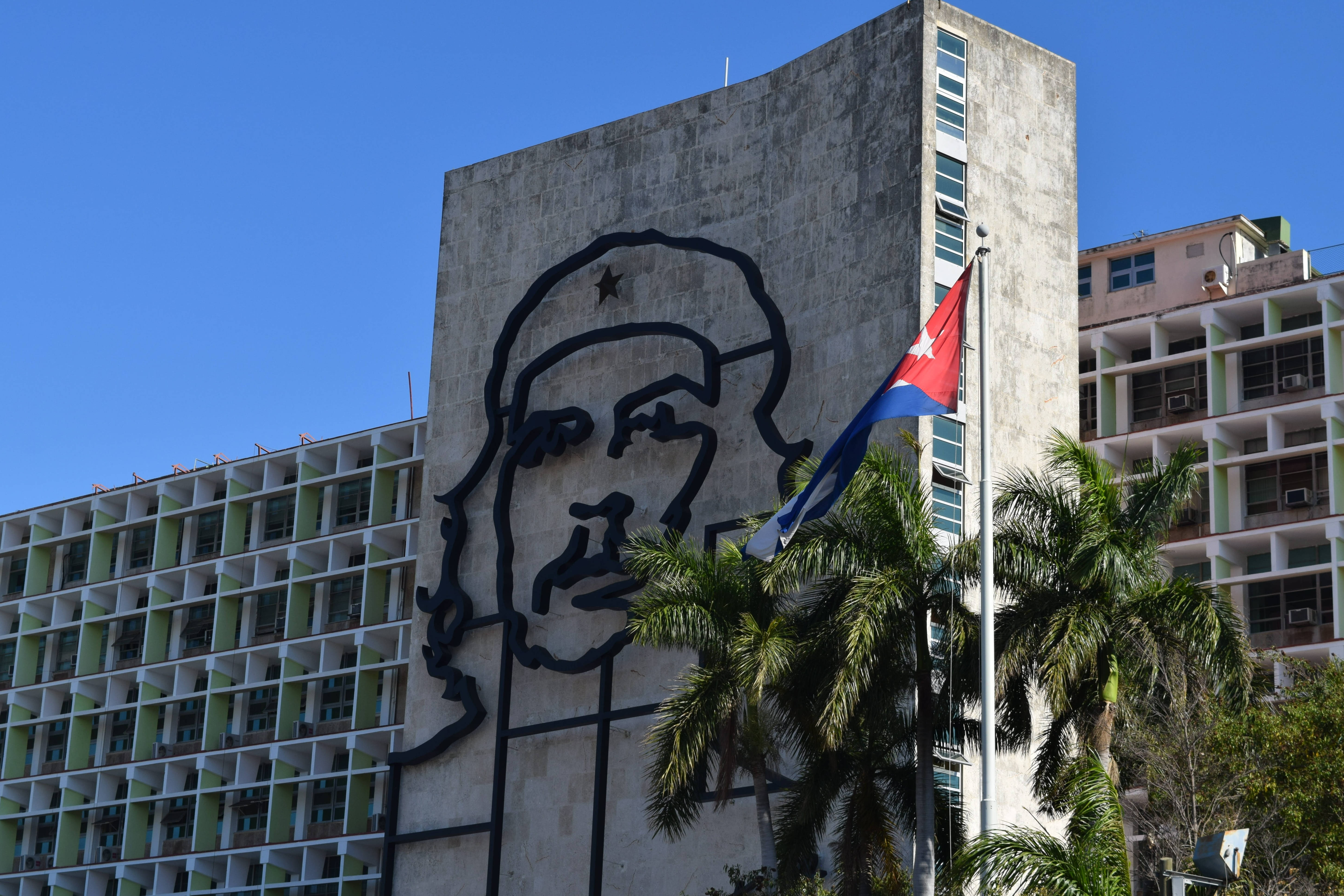 Cuban 桌布 壁紙 — 一個巨大的黑色切·格瓦拉臉部輪廓主導著一座混凝土建築，背景是晴朗的藍天、棕櫚樹和附近飄揚的古巴國旗。
