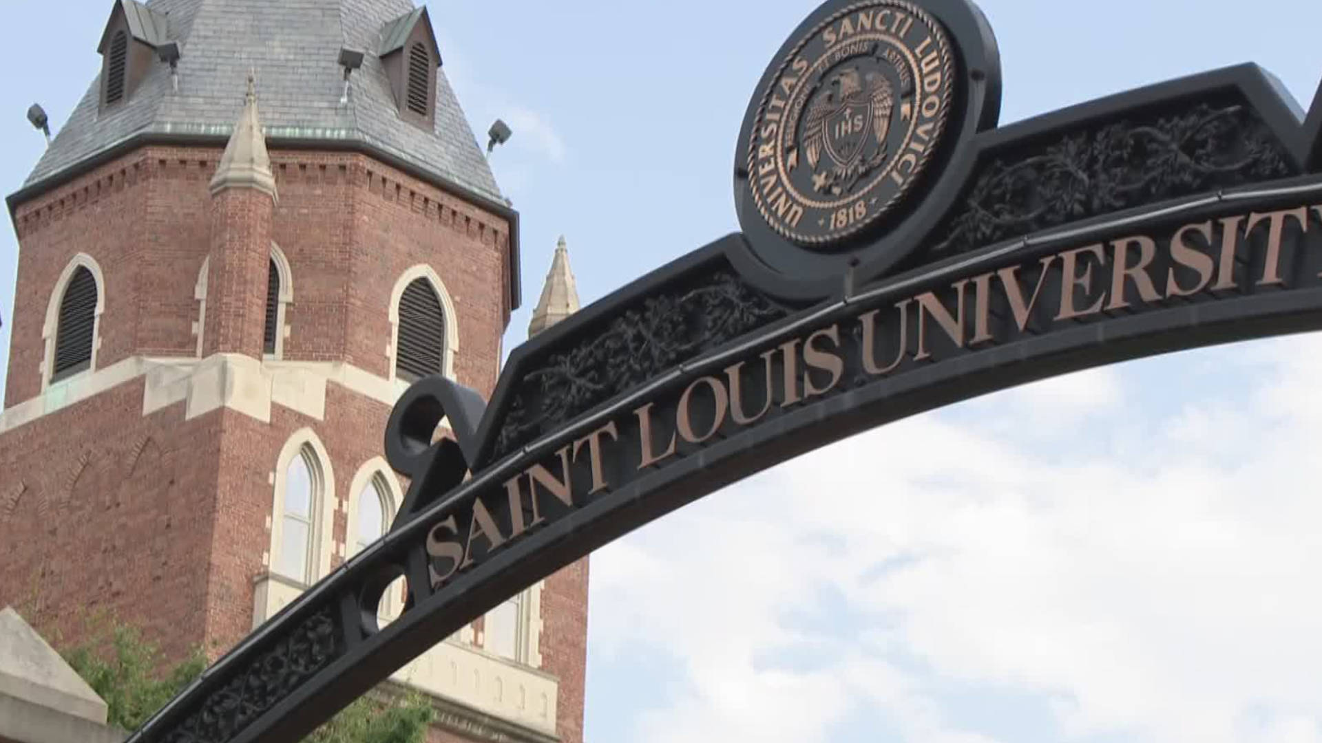 建築 桌布 壁紙 — 一個巨大的黑色鍛鐵拱門，上面寫著「SAINT LOUIS UNIVERSITY」，矗立在歷史悠久的紅磚哥德式塔樓前，天空部分多雲。