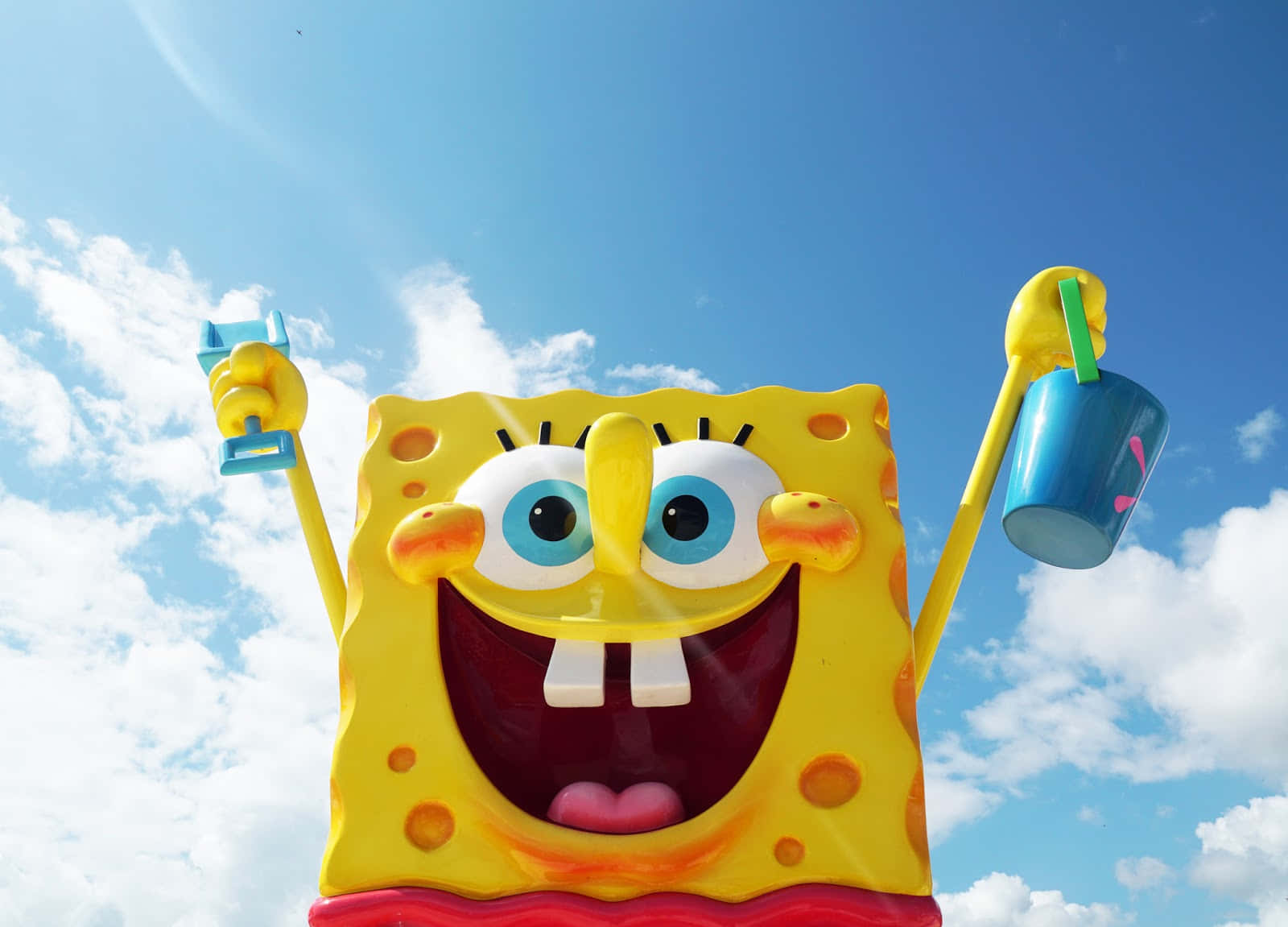 nickelodeon 桌布 壁紙 — 一個巨大開心的黃色海綿角色雕像，拿著一個藍色杯子和一個藍色小物件，背景是明亮的藍天和蓬鬆的白雲。